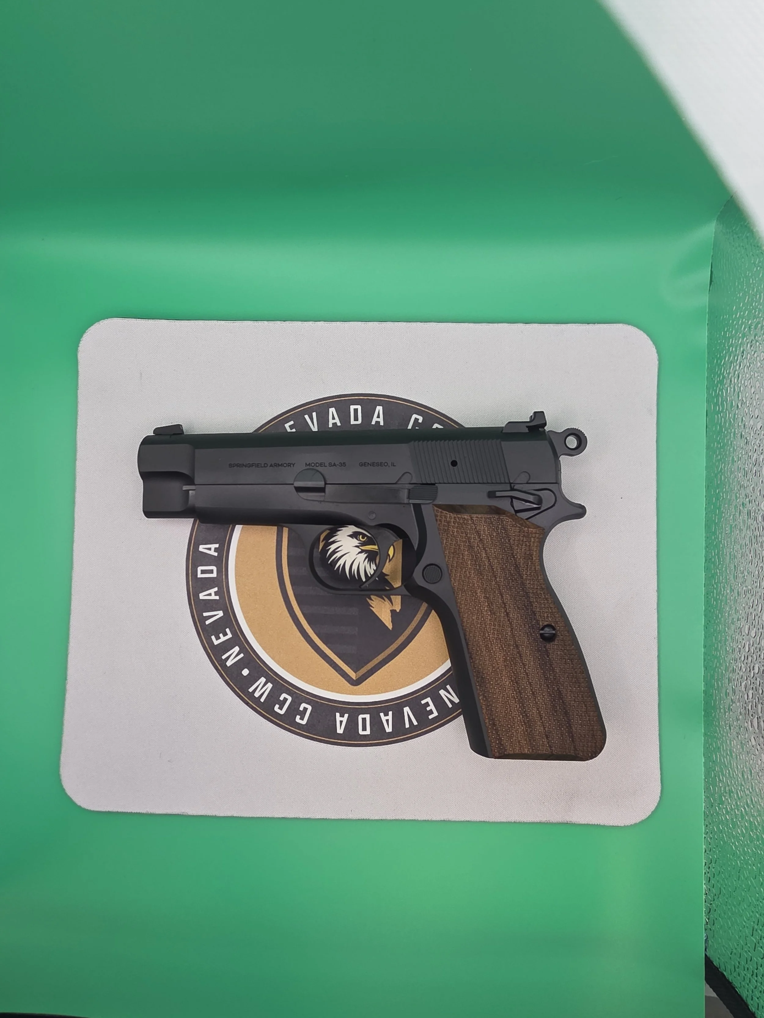 Close-up of a black handgun with wooden grips, placed on a white surface with a Nevada CCW badge behind it, inside a green box.