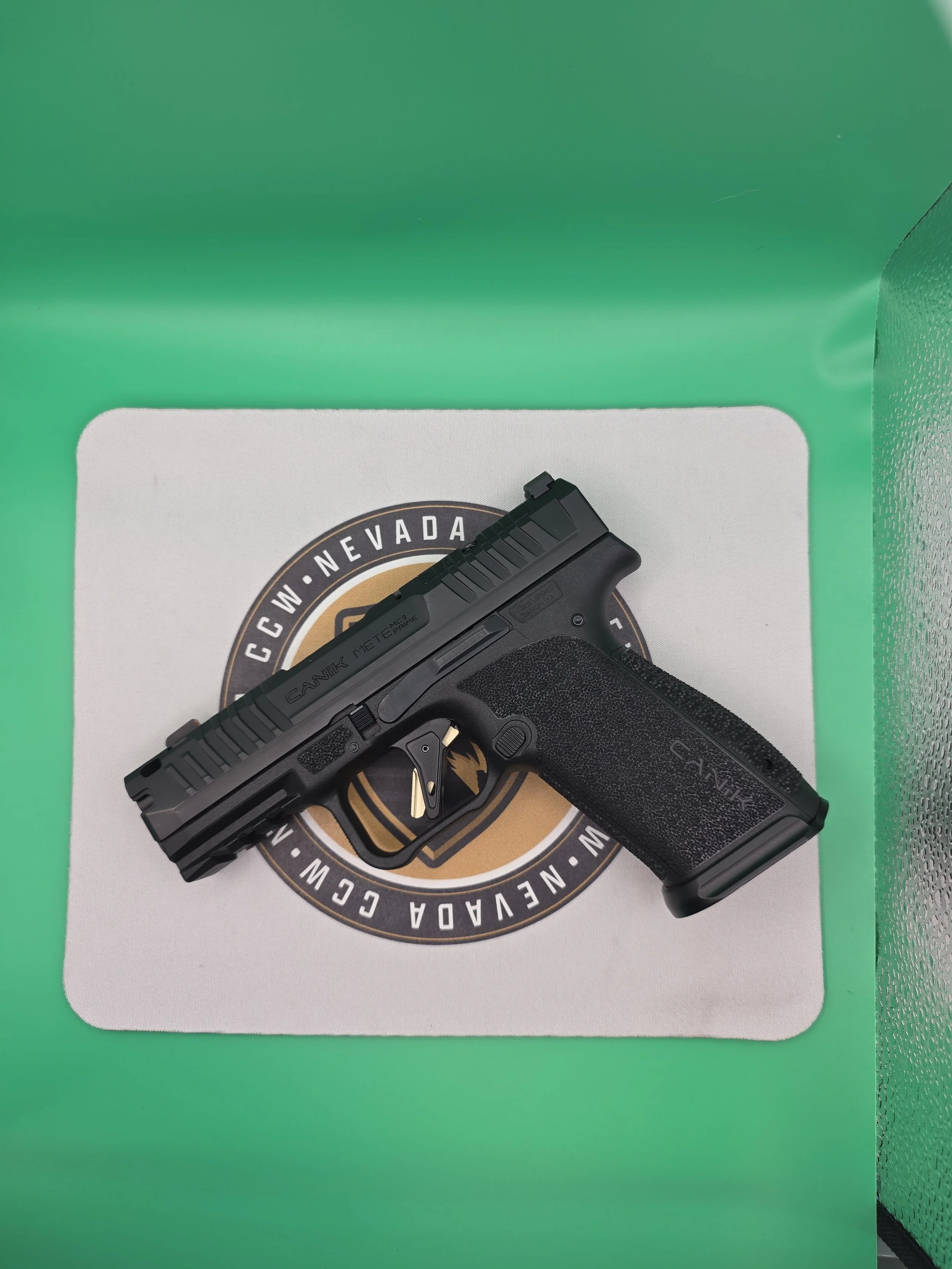 A black handgun with a textured grip placed on a white surface with a circular logo surrounding the text 'W. Nevada' and other partially visible words, with a green border and a textured black side panel.