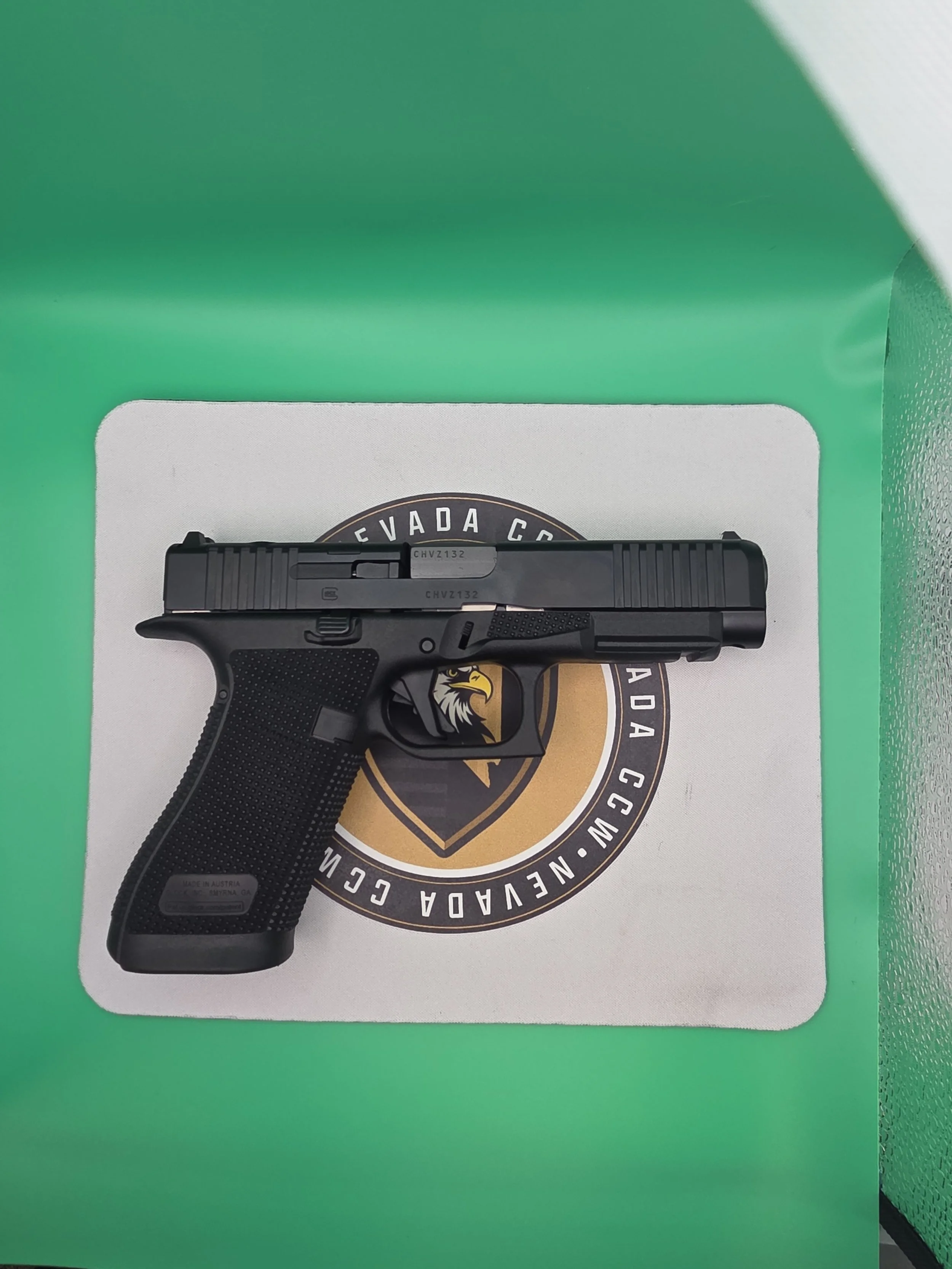 A black semi-automatic pistol placed on a white surface with a circular logo of Nevada County, California underneath it. The background is a green surface with some textured areas.