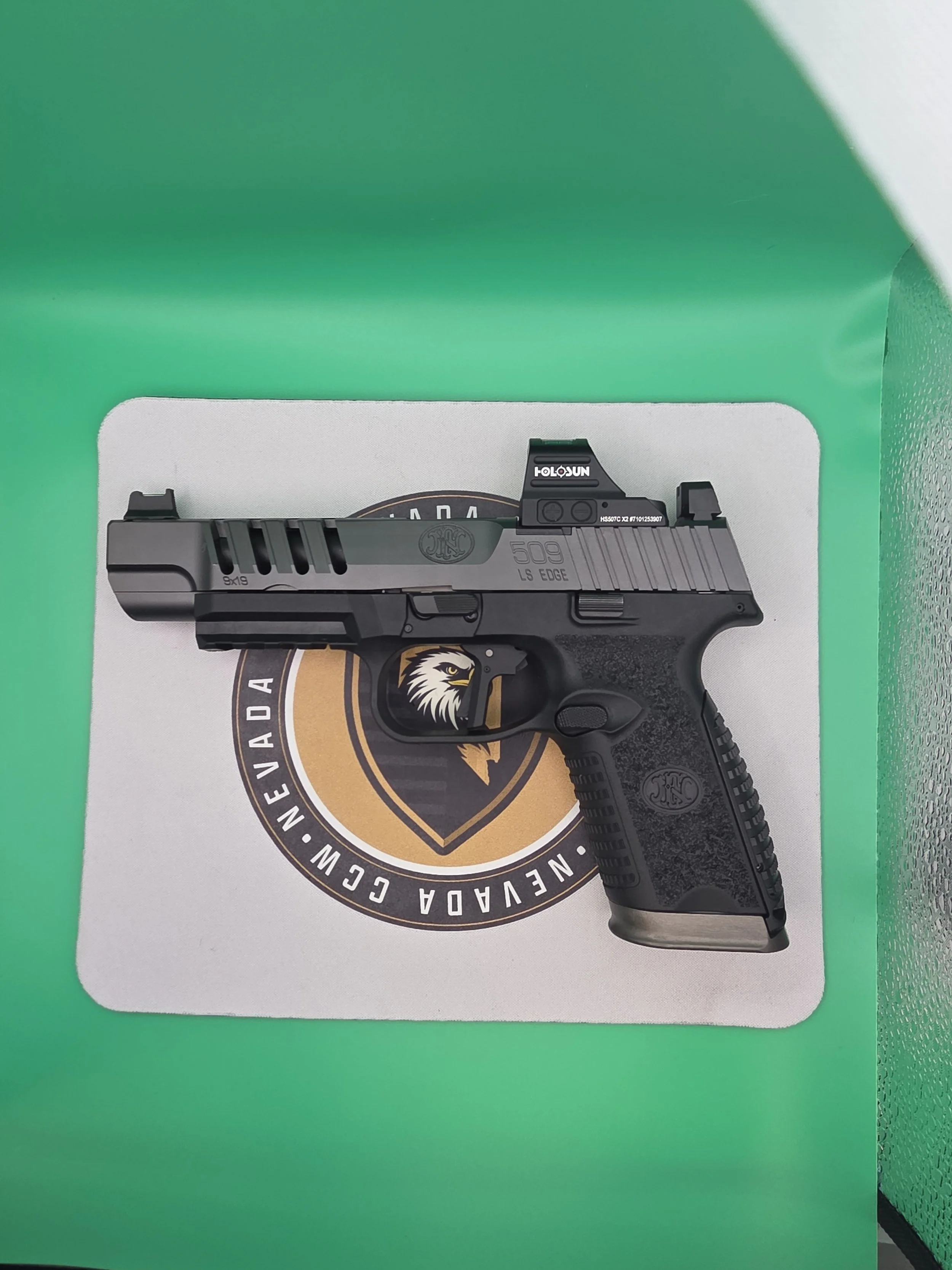 A black handgun with a textured grip, mounted with a holographic sight labeled 'HOLOSUN,' resting on a white surface with a logo surrounded by a circular text.