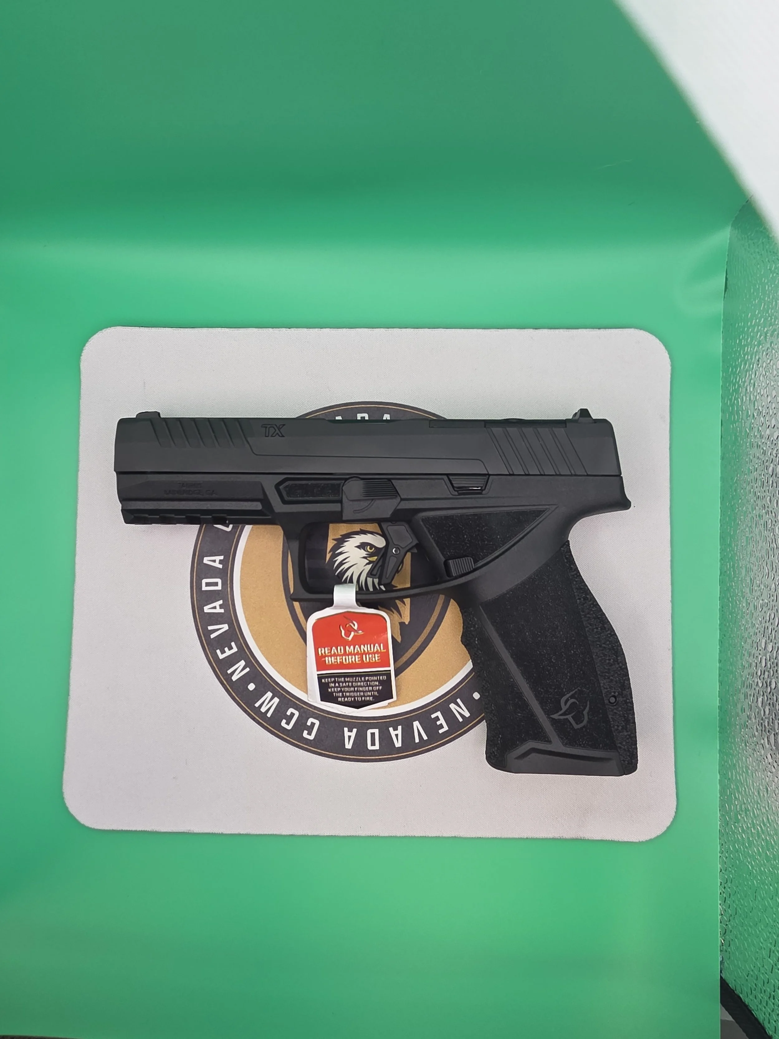 Black semi-automatic handgun with textured grip resting on a printed sheet with circular logo, against a green background.