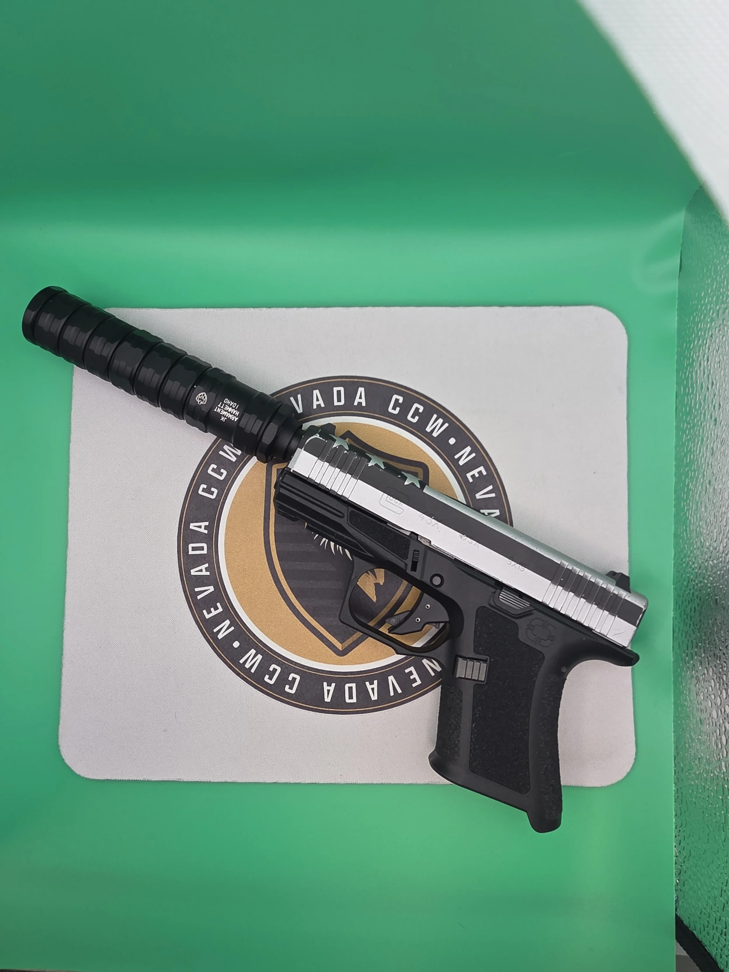 A handgun with a wooden grip resting on a white box that has a circular logo with black, gold, and green colors, surrounded by the words "Nevada CCW." The box is on a green surface with a green wall in the background.