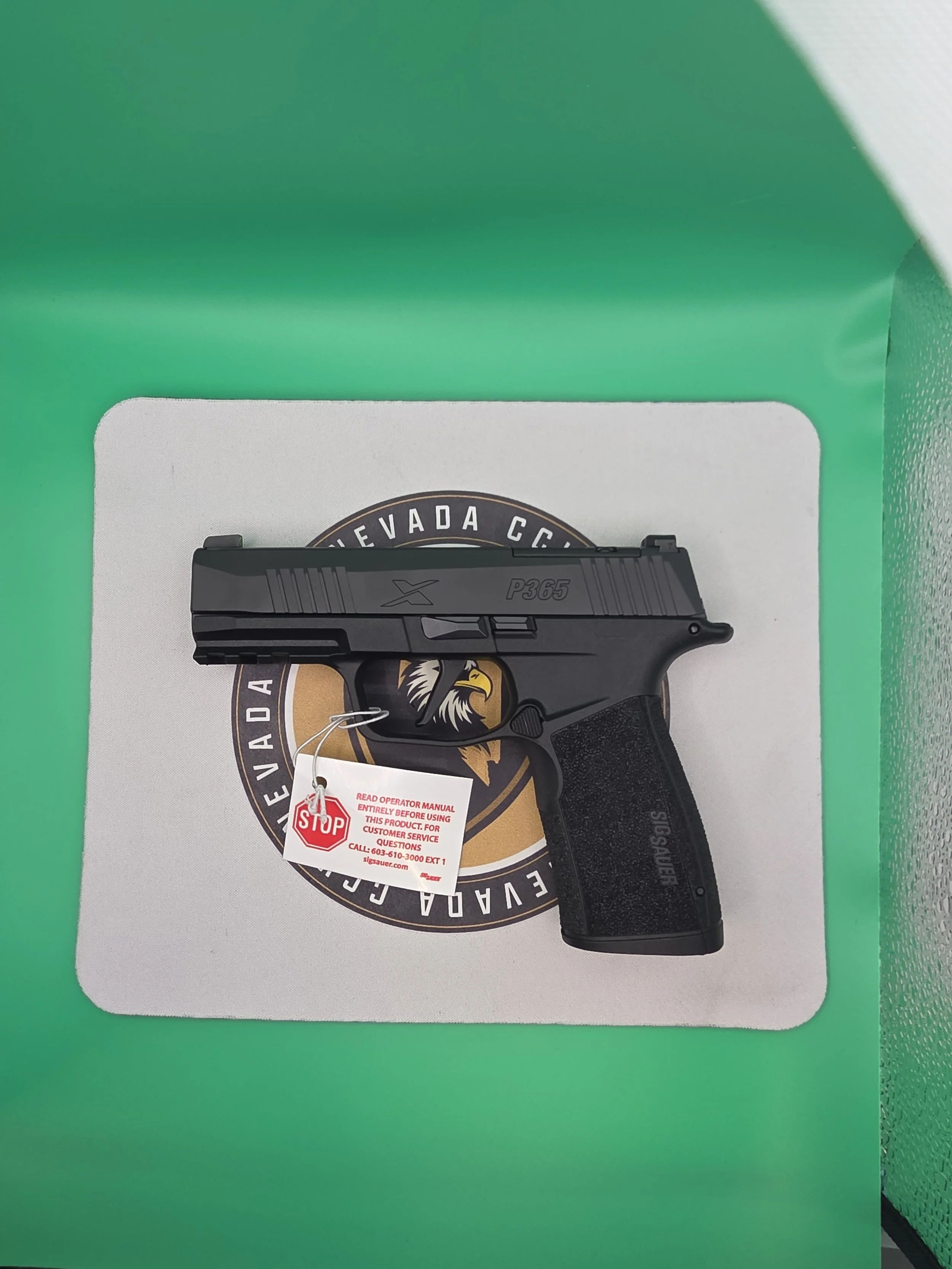 A black Sig Sauer P365 handgun with a textured grip displayed on a white surface, with a warning tag attached, against a background of green and white patterned surfaces.