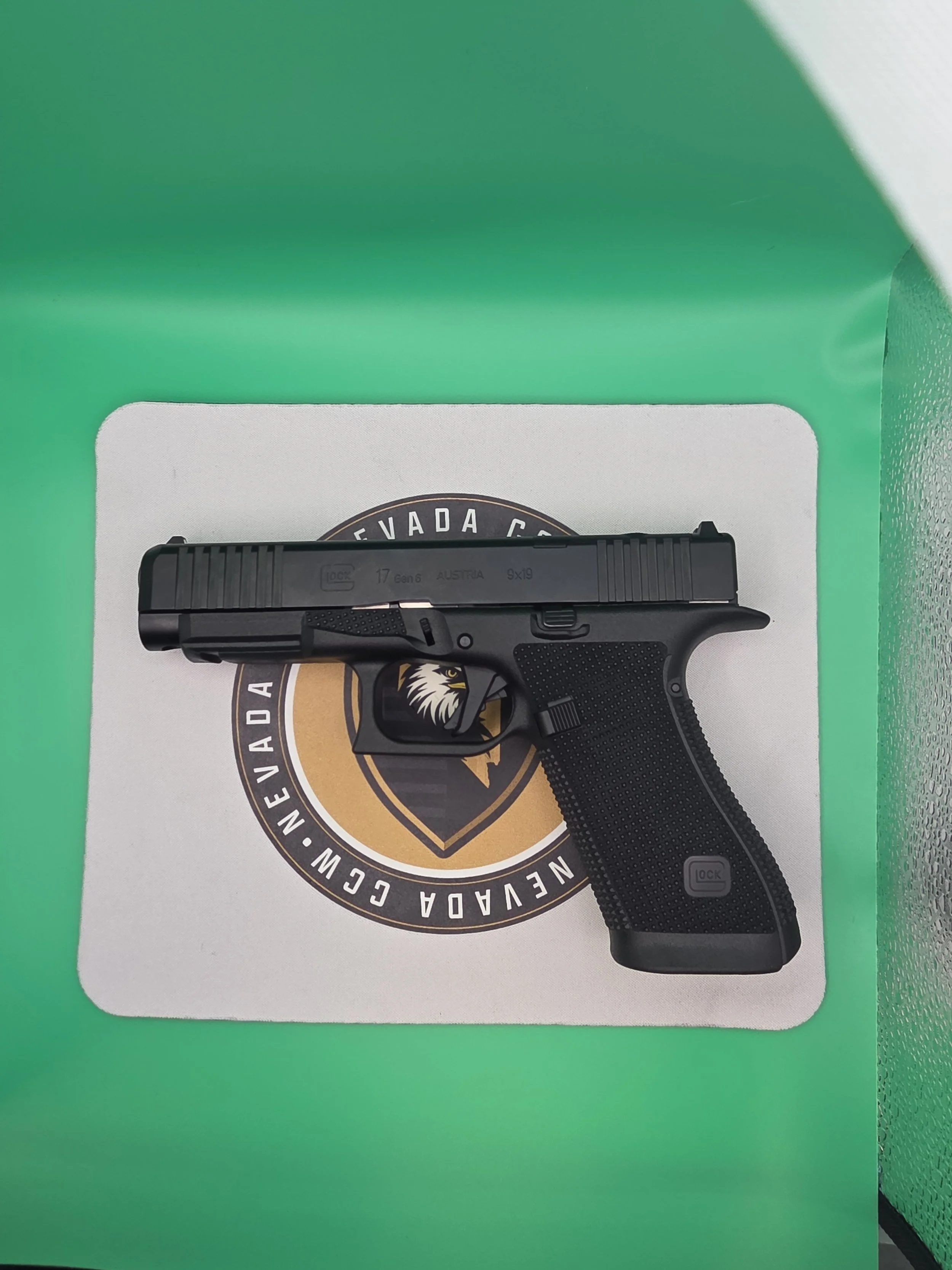 A black Glock handgun resting on a white surface with a circular logo, in front of a green background.