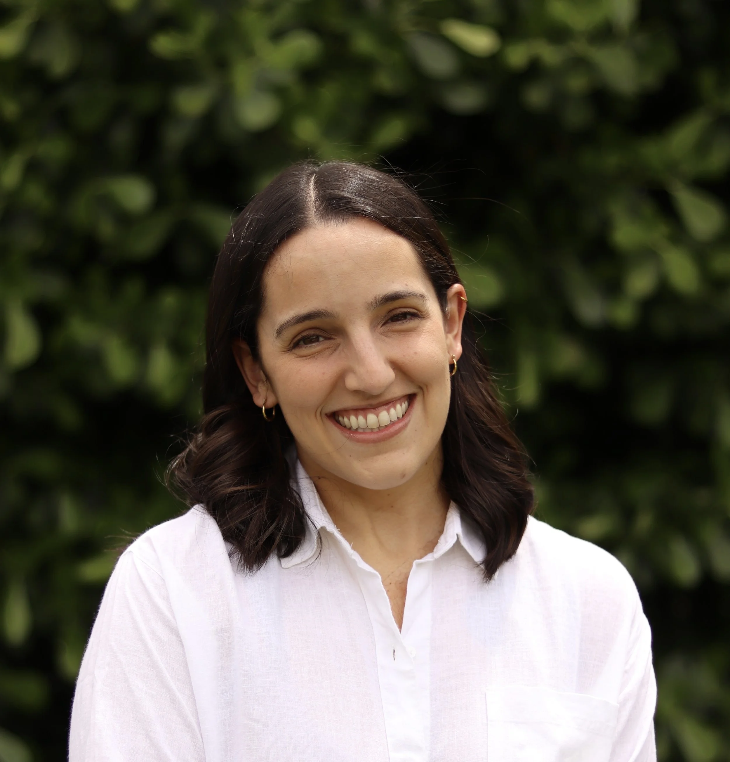 Close-up of a smiling woman with long dark hair, wearing a white shirt, outdoors with blurred green foliage in the background.