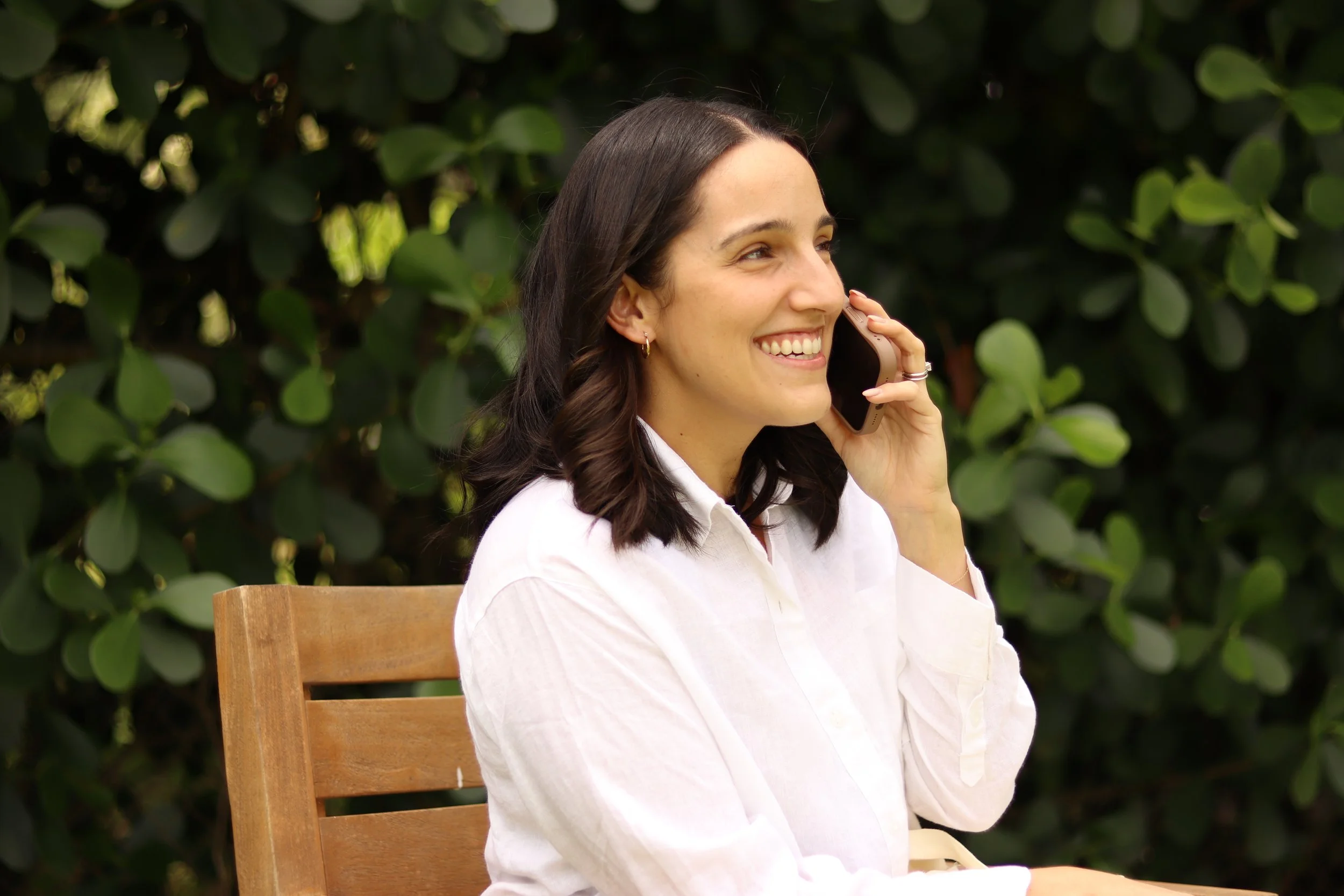 A woman smiling and talking on a cellphone while sitting outdoors on a wooden chair, with green leafy bushes in the background.