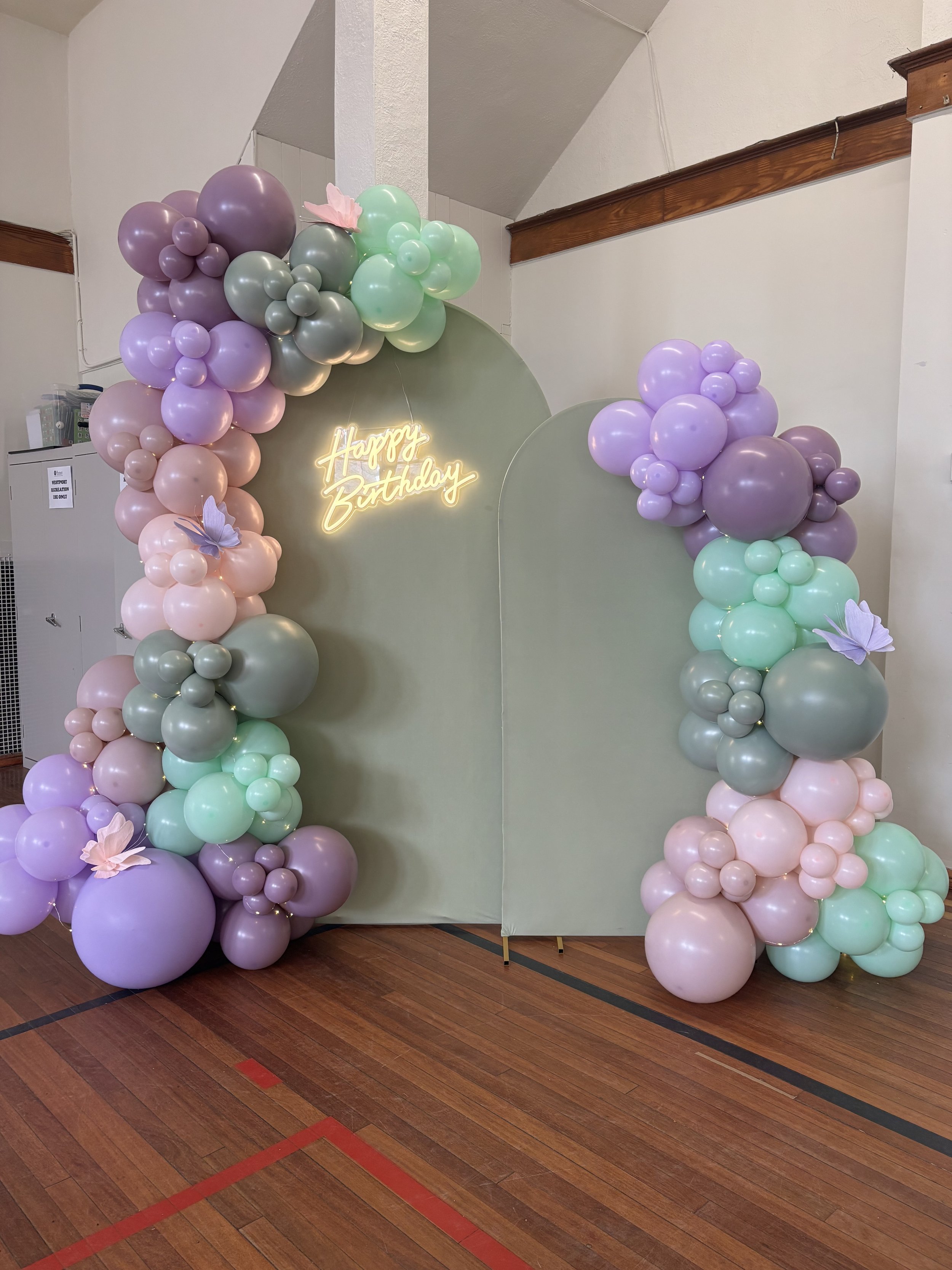 Decorative balloon arch with pastel colors around a room divider, featuring a neon sign that says 'Happy Birthday' and paper butterflies.