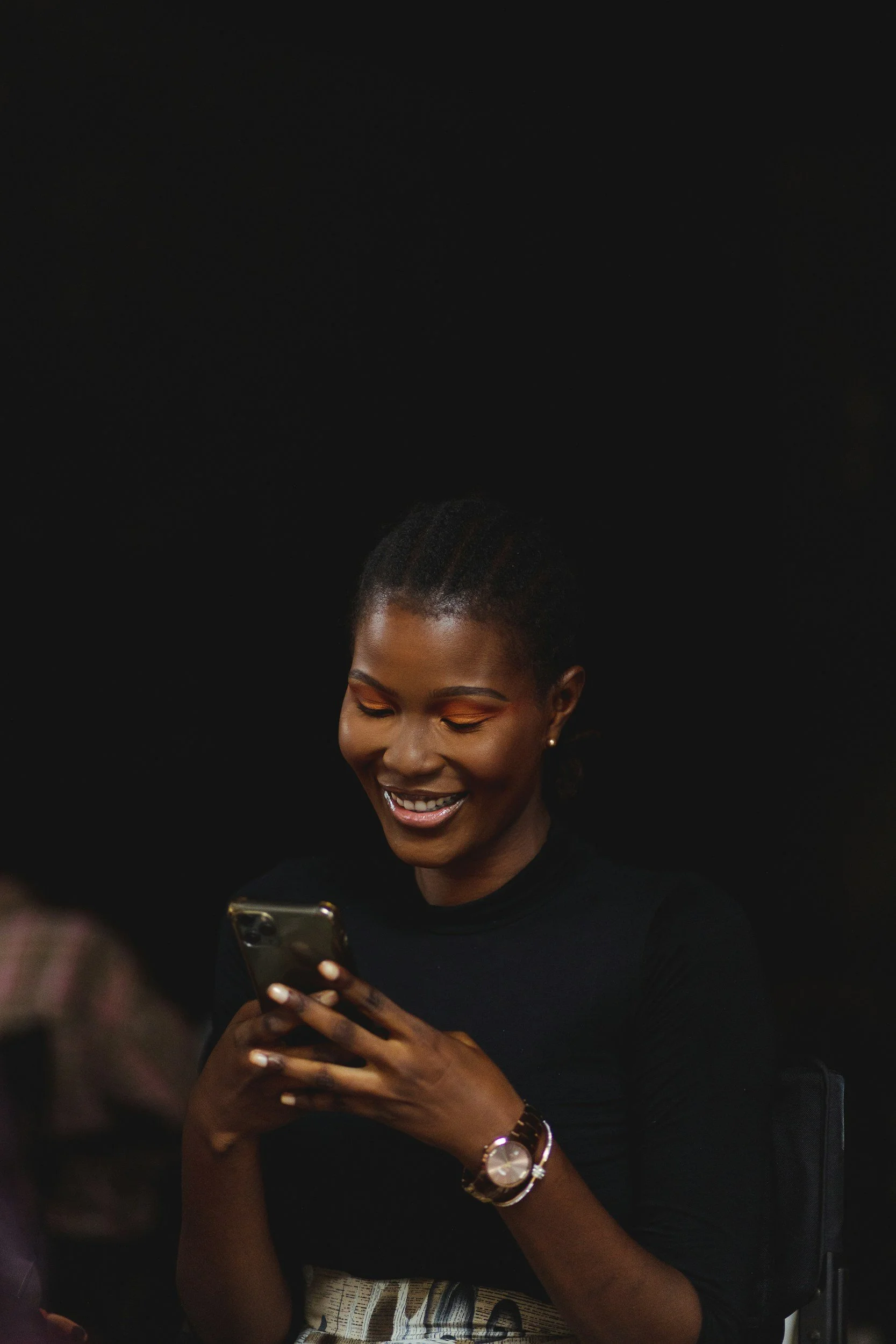A woman with dark skin and braided hair smiling while looking at her phone against a black background.