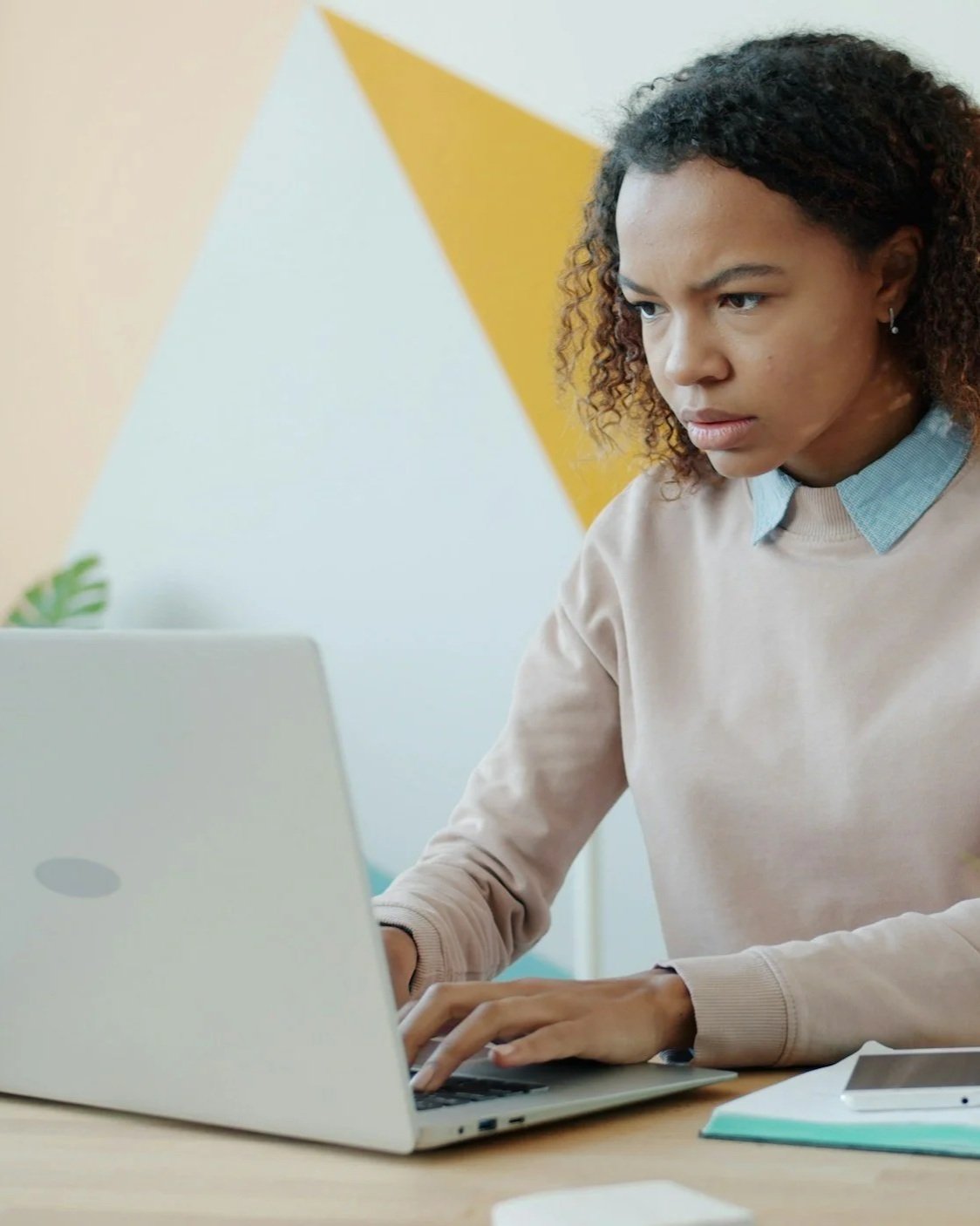 A woman with curly hair working on a laptop at a desk, focused and serious.