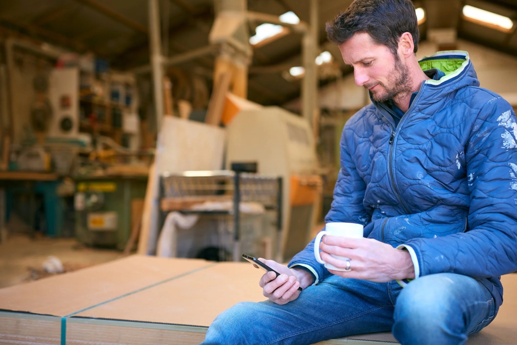 A man sitting on a wooden table in a workshop or warehouse, holding a white mug in one hand and a smartphone in the other, wearing a blue quilted jacket and blue jeans, with shelves of tools and equipment in the background.
