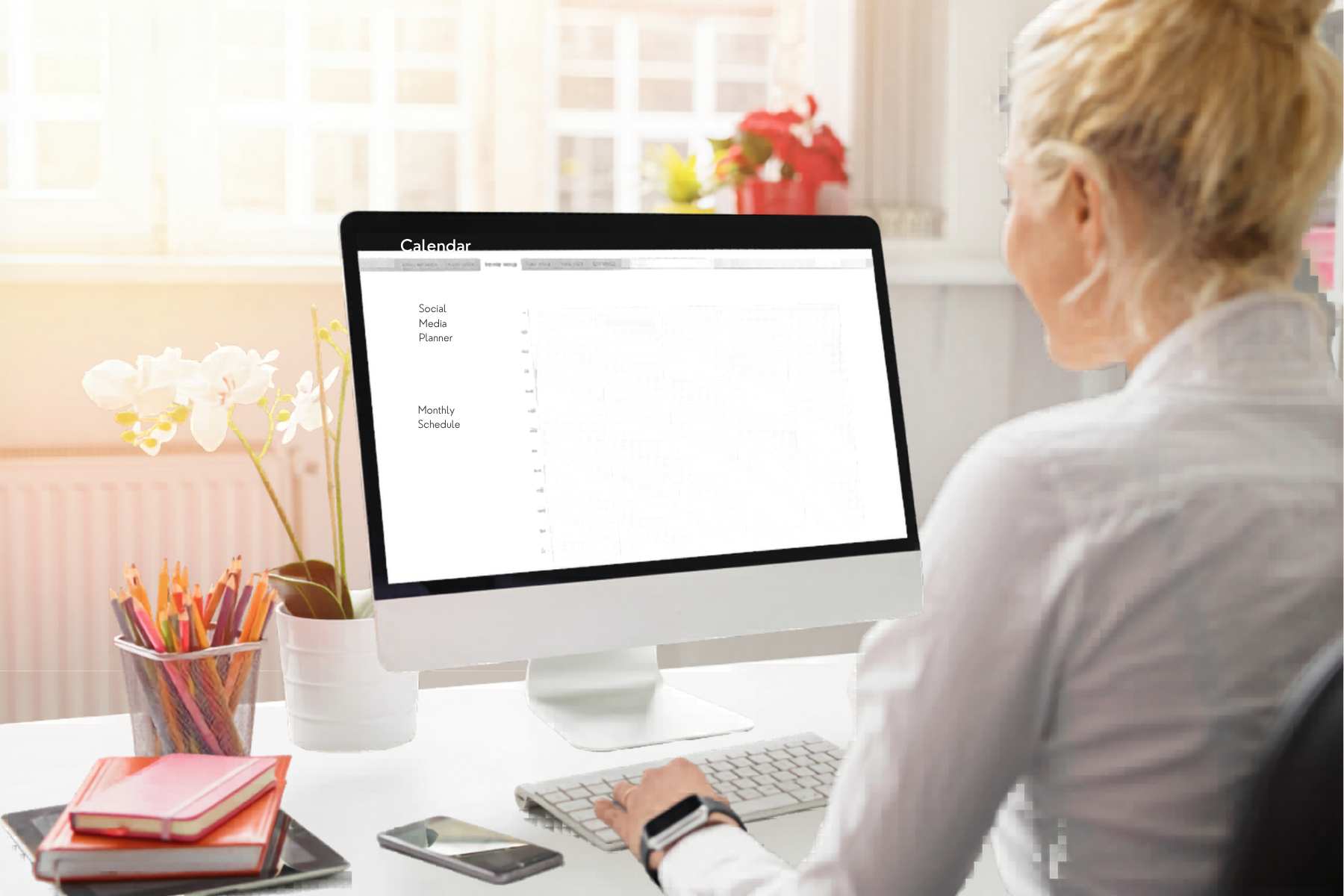 A woman working at a white desk in front of a computer monitor displaying a planner. On the desk, there are notebooks, a smartphone, a tablet, a glass container with colored pencils, a white pot with white orchids, and a pink flower pot with red flowers in the background. The room is well-lit with natural sunlight from a window.