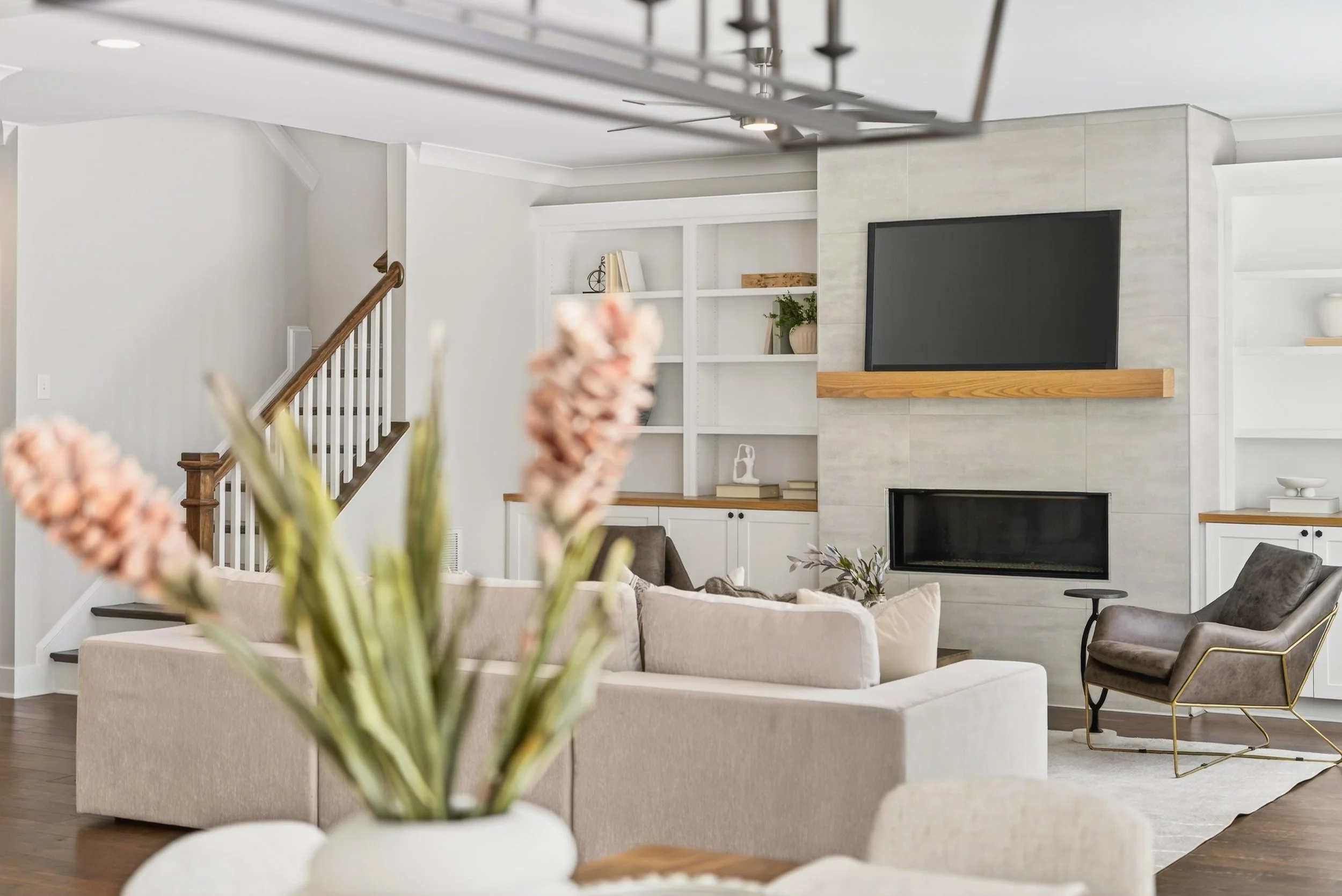 Living room with a beige couch, dark gray armchair, and white built-in shelves around a fireplace with a large flat-screen TV, decorated with books and plants.