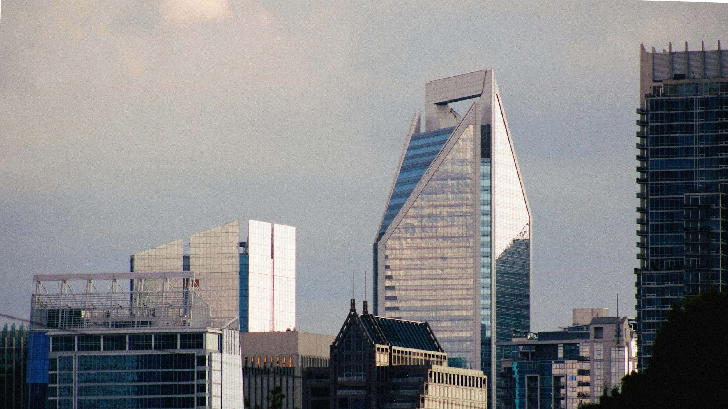 A city skyline featuring modern skyscrapers with glass exteriors, including one tall, pointed building with a triangular shape, under a cloudy sky.