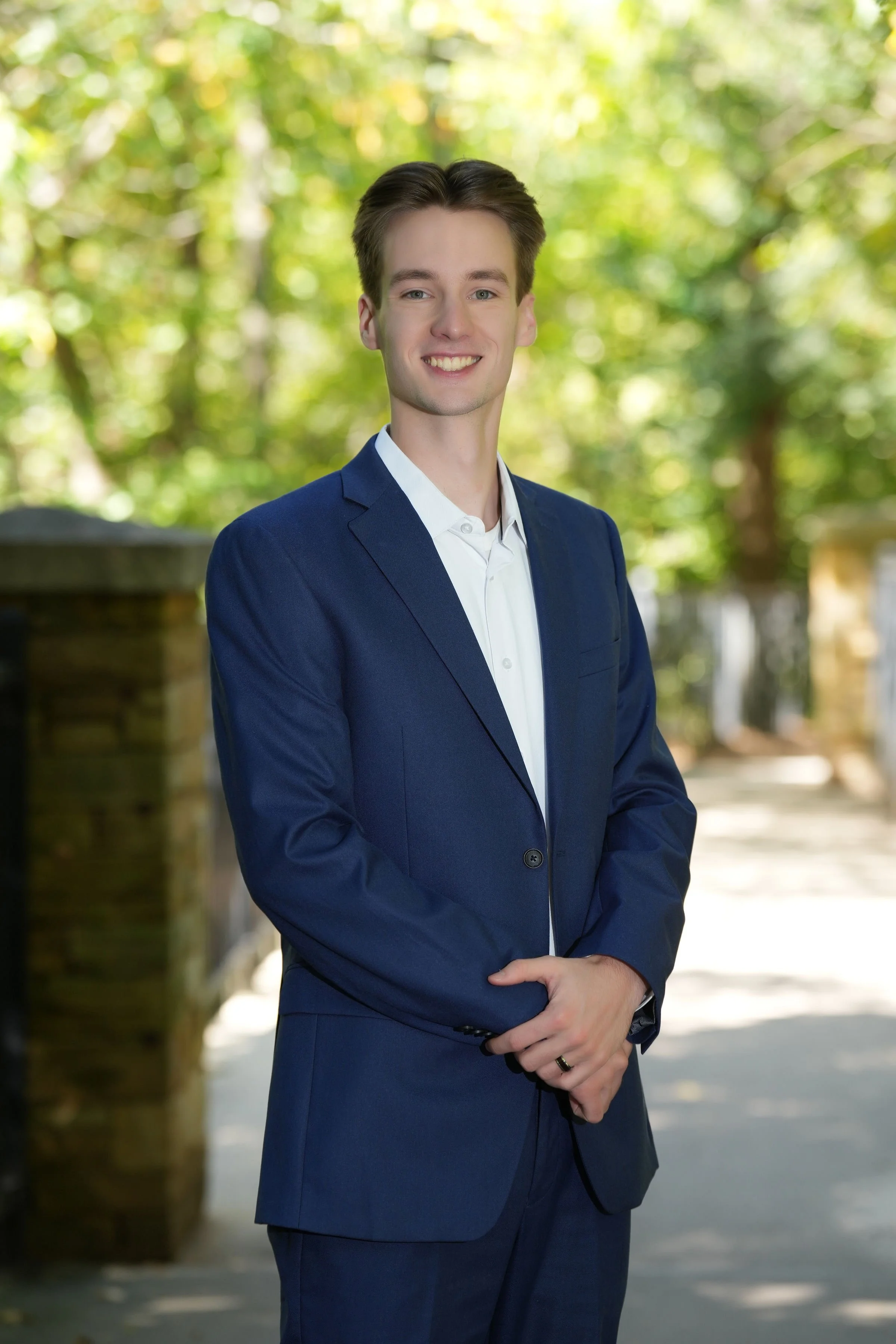 Head and shoulders of a young man with brown hair in a blue suit and white shirt, smiling, standing outdoors on a sunny day with green trees in the background.