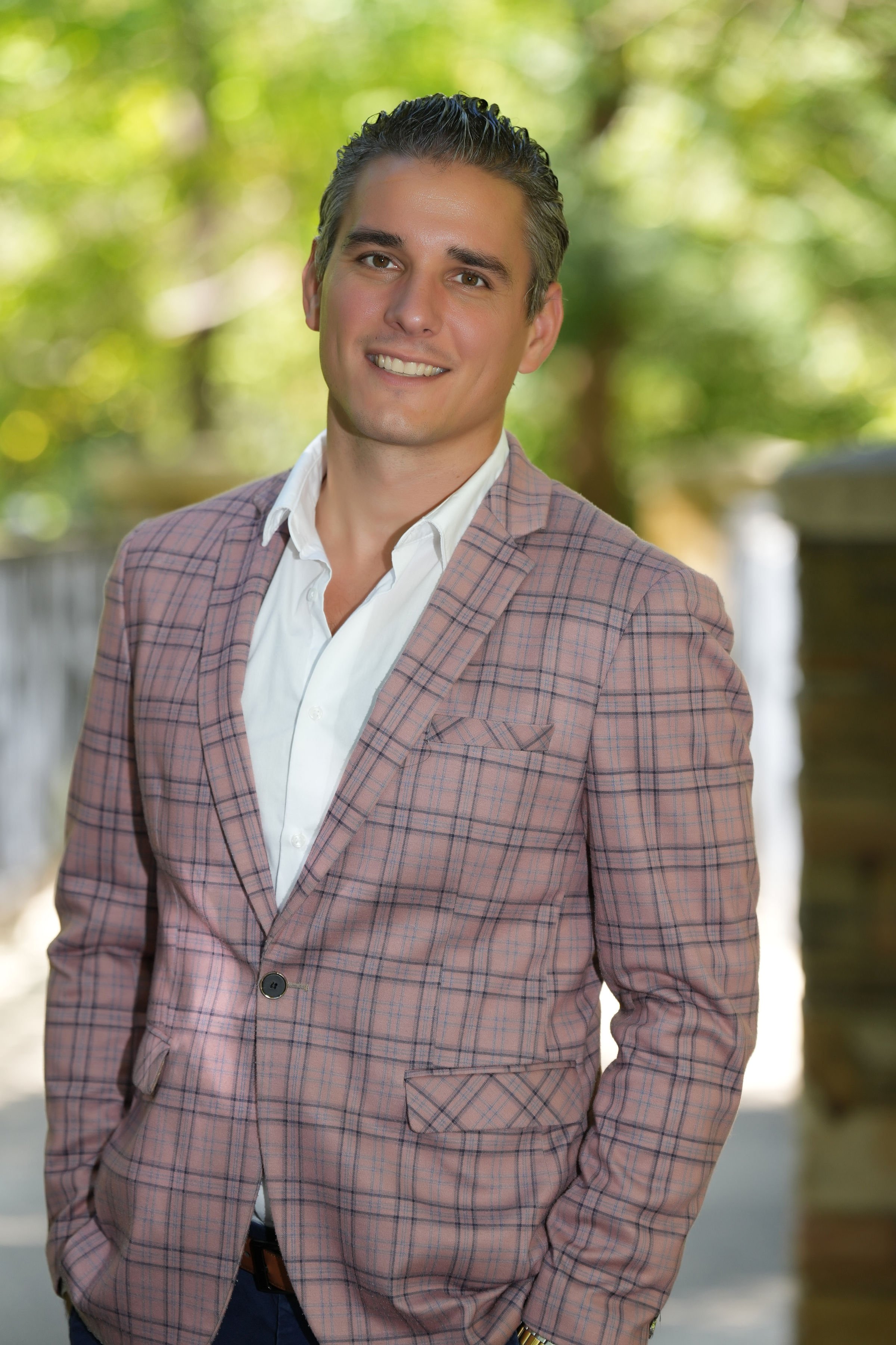 Headshot of a smiling man wearing a light checkered blazer and white shirt, outdoors with greenery in the background.