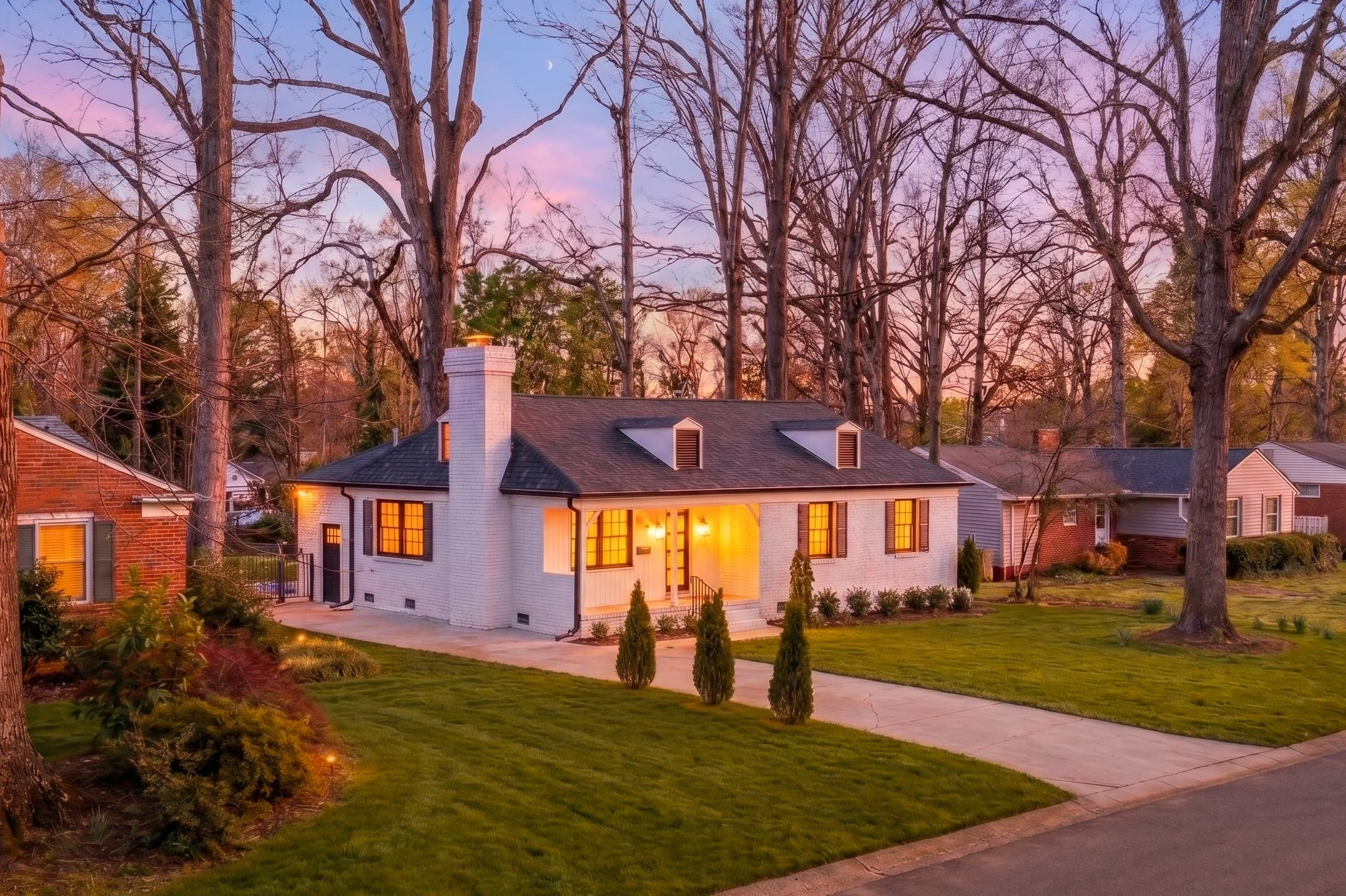 A cozy white house with warm interior lighting, surrounded by a well-maintained lawn and small trees, during sunset with leafless trees and a moon visible in the sky.