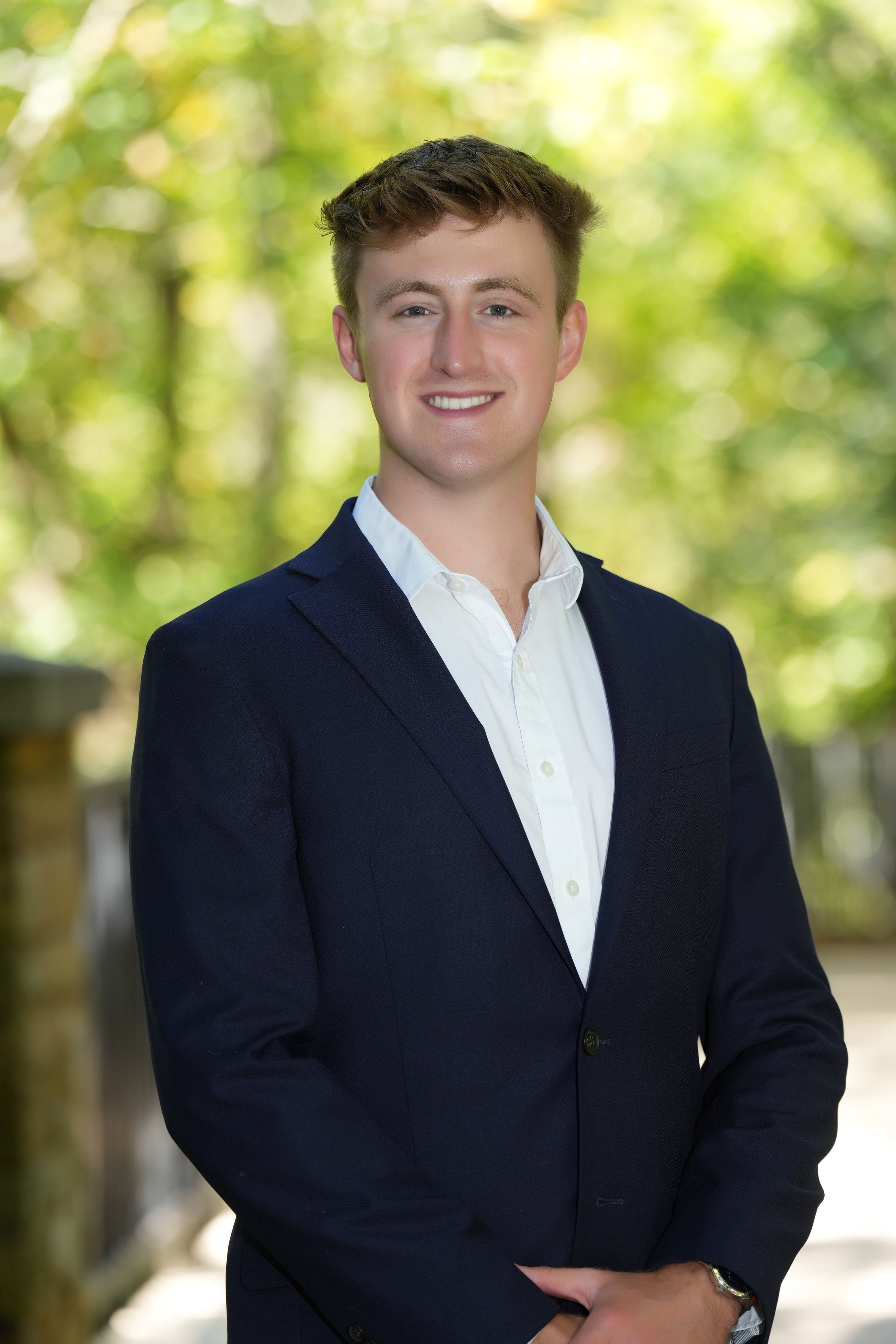 A young man with short brown hair and blue eyes wearing a dark blue suit and white shirt, standing outdoors with a blurred green background.