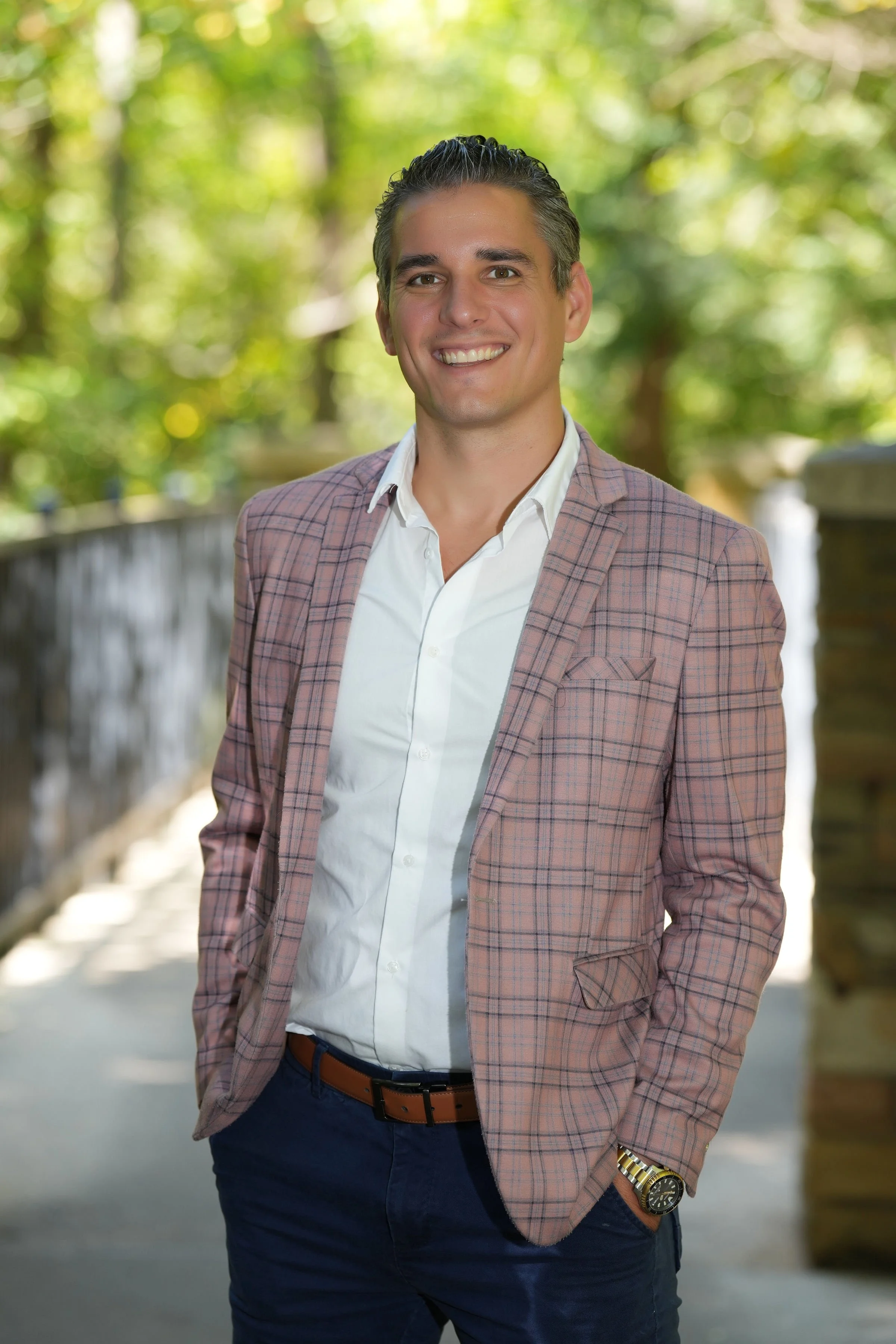 Smiling man in a tan plaid blazer, white shirt, and blue pants, standing outdoors on a sunny day with green trees in the background.