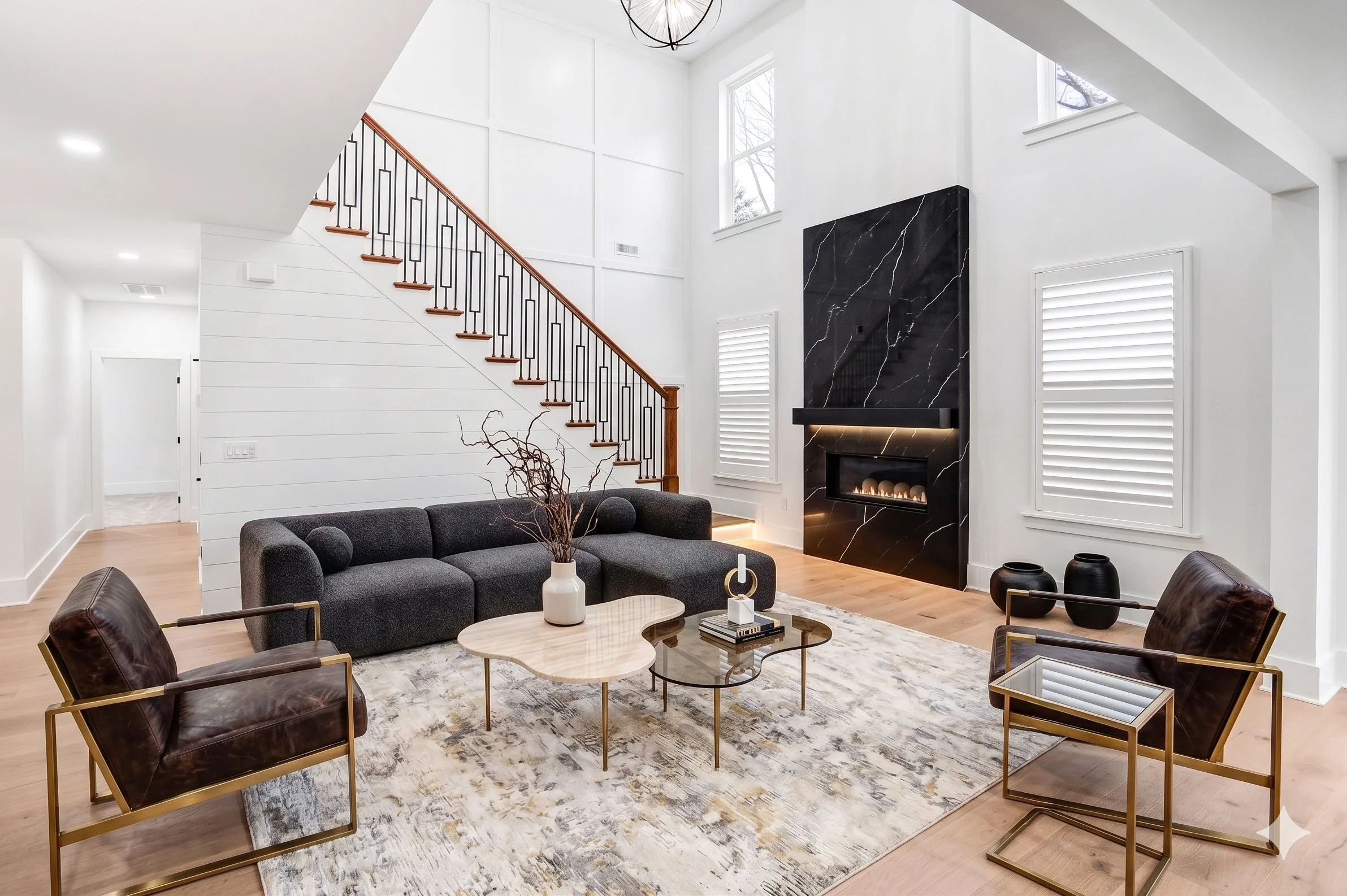 Modern living room with white walls, high ceilings, a black marble fireplace, wooden staircase with black metal railing, large windows, and contemporary furniture including a gray sofa, leather chairs, and coffee tables.