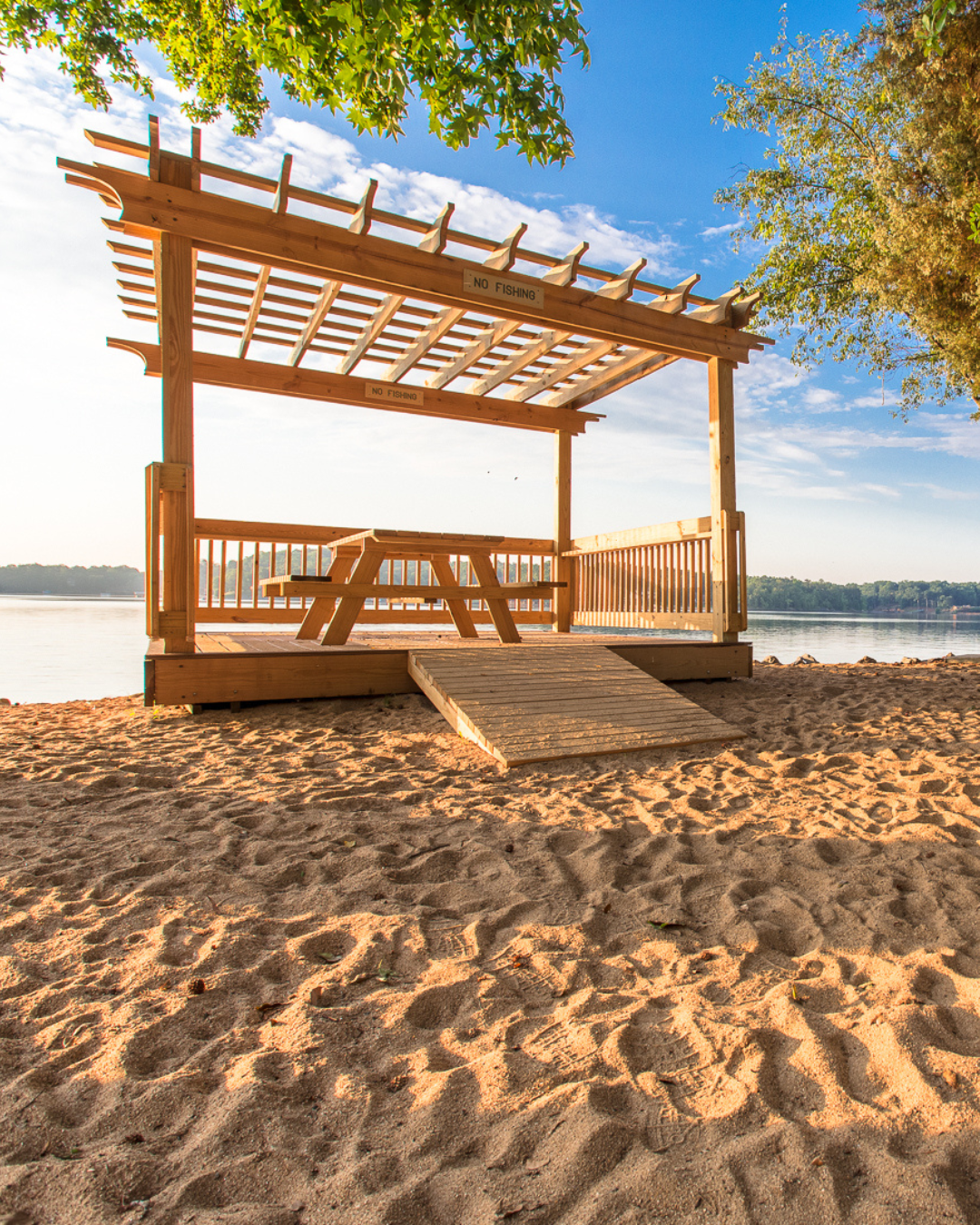 Wooden pavilion with a picnic table and a ramp on sandy beach beside a body of water, with trees and blue sky in the background.
