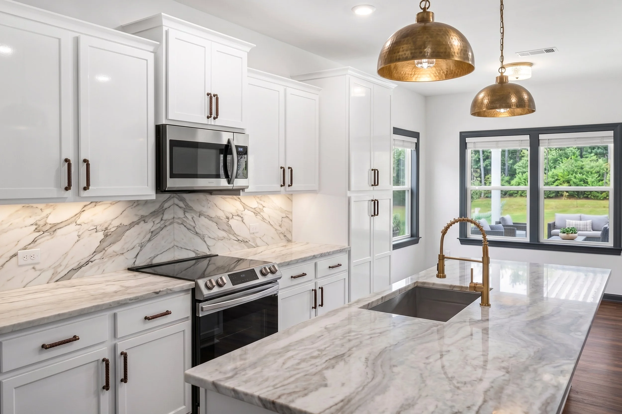 Modern kitchen with white cabinets, marble countertops and backsplash, stainless steel microwave, black stove, gold pendant lights, and large windows showing a green outdoor view.