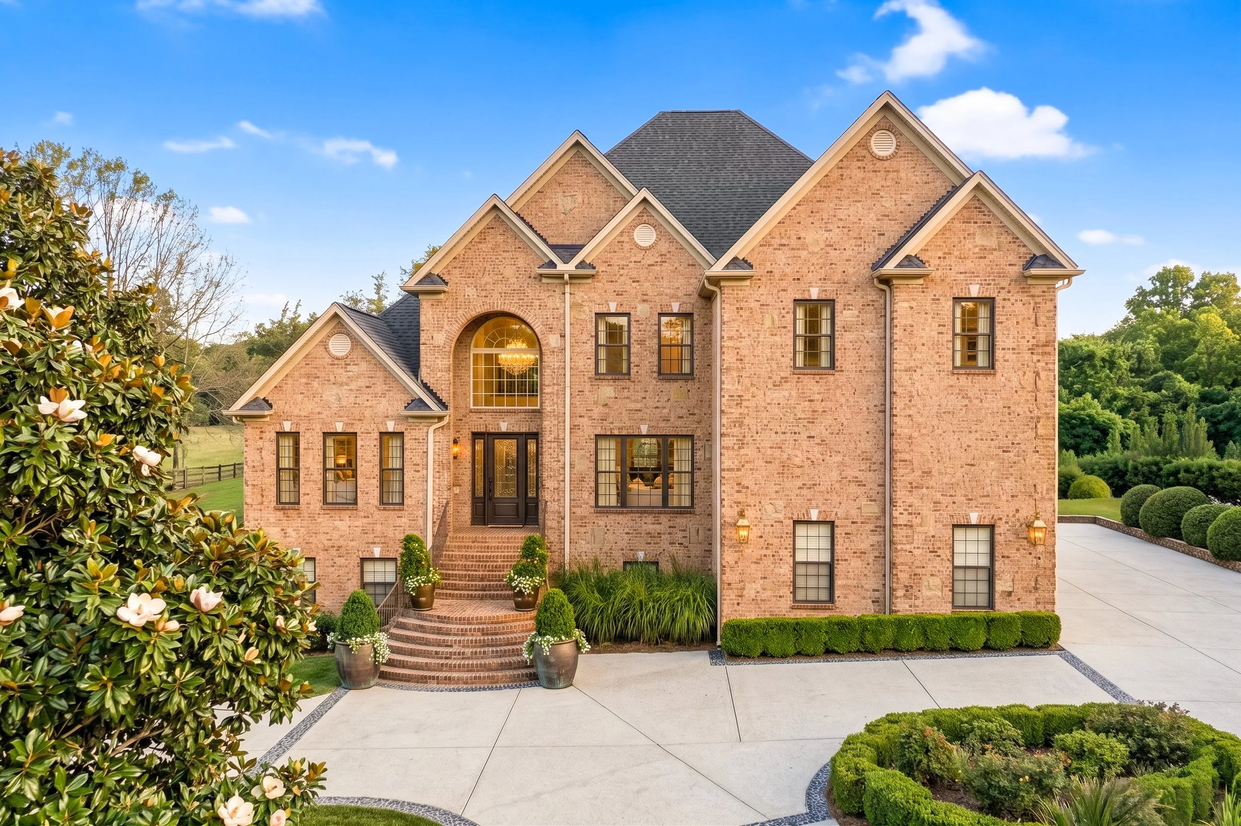 A large brick house with multiple gabled roofs, tall windows, and a grand arched window with a chandelier inside. The house is surrounded by well-manicured bushes, potted plants, and a paved driveway under a clear blue sky.