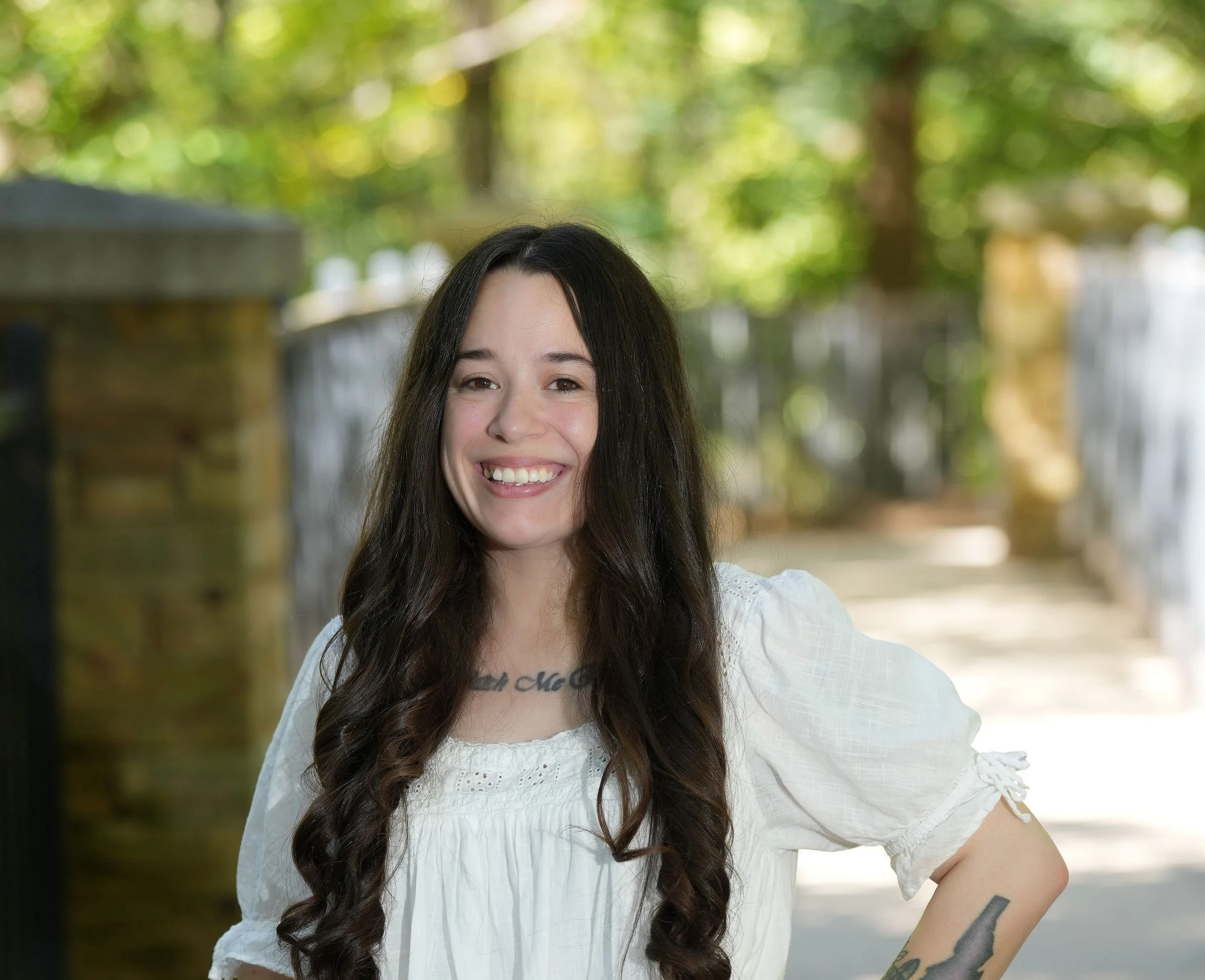 A young woman with long dark hair smiling outdoors, wearing a white eyelet top, with a tattoo on her forearm, in front of a blurred background of trees and a wooden fence.