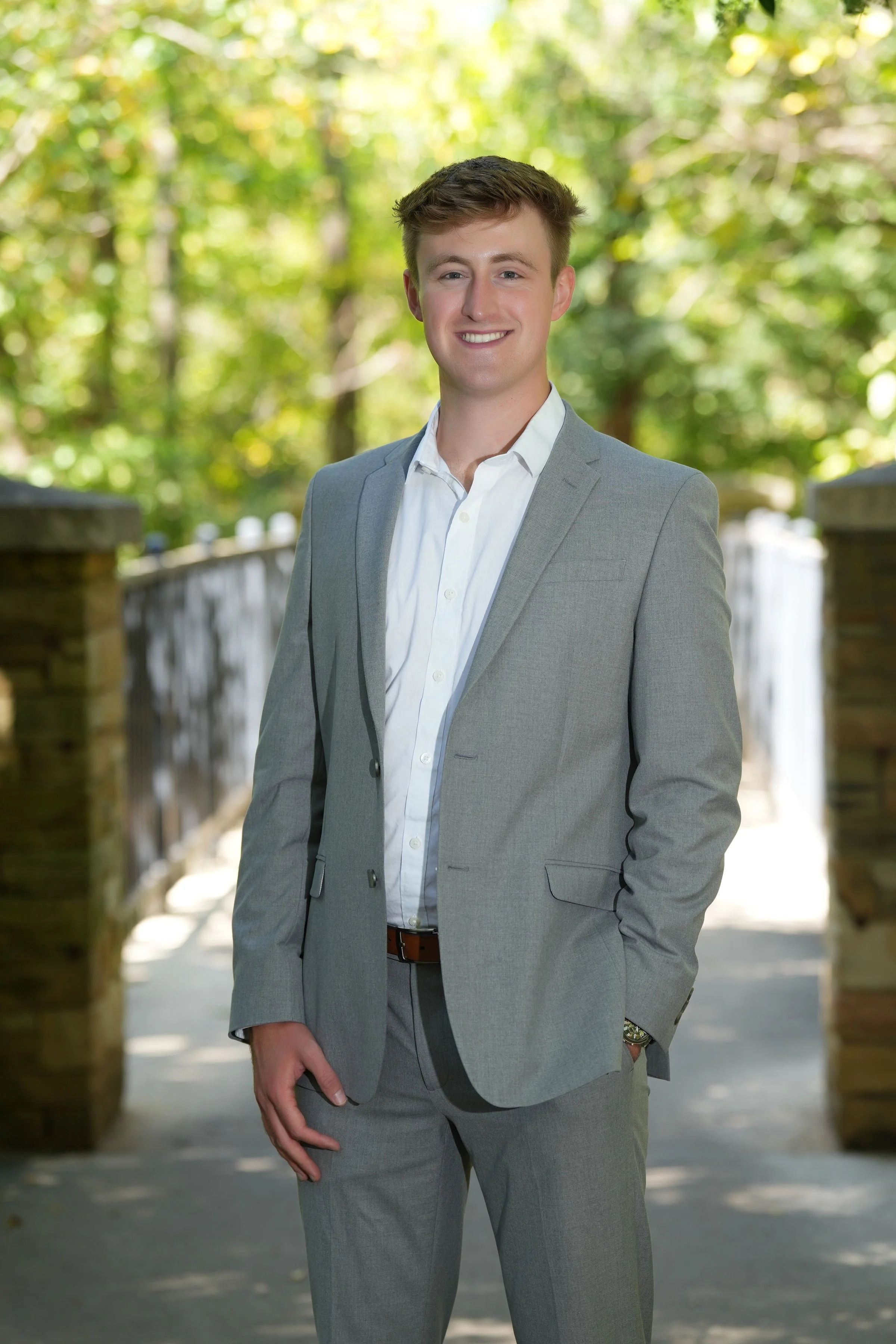 A young man in a gray suit with a white shirt standing outdoors on a pathway, with trees and a wooden fence in the background, smiling at the camera.