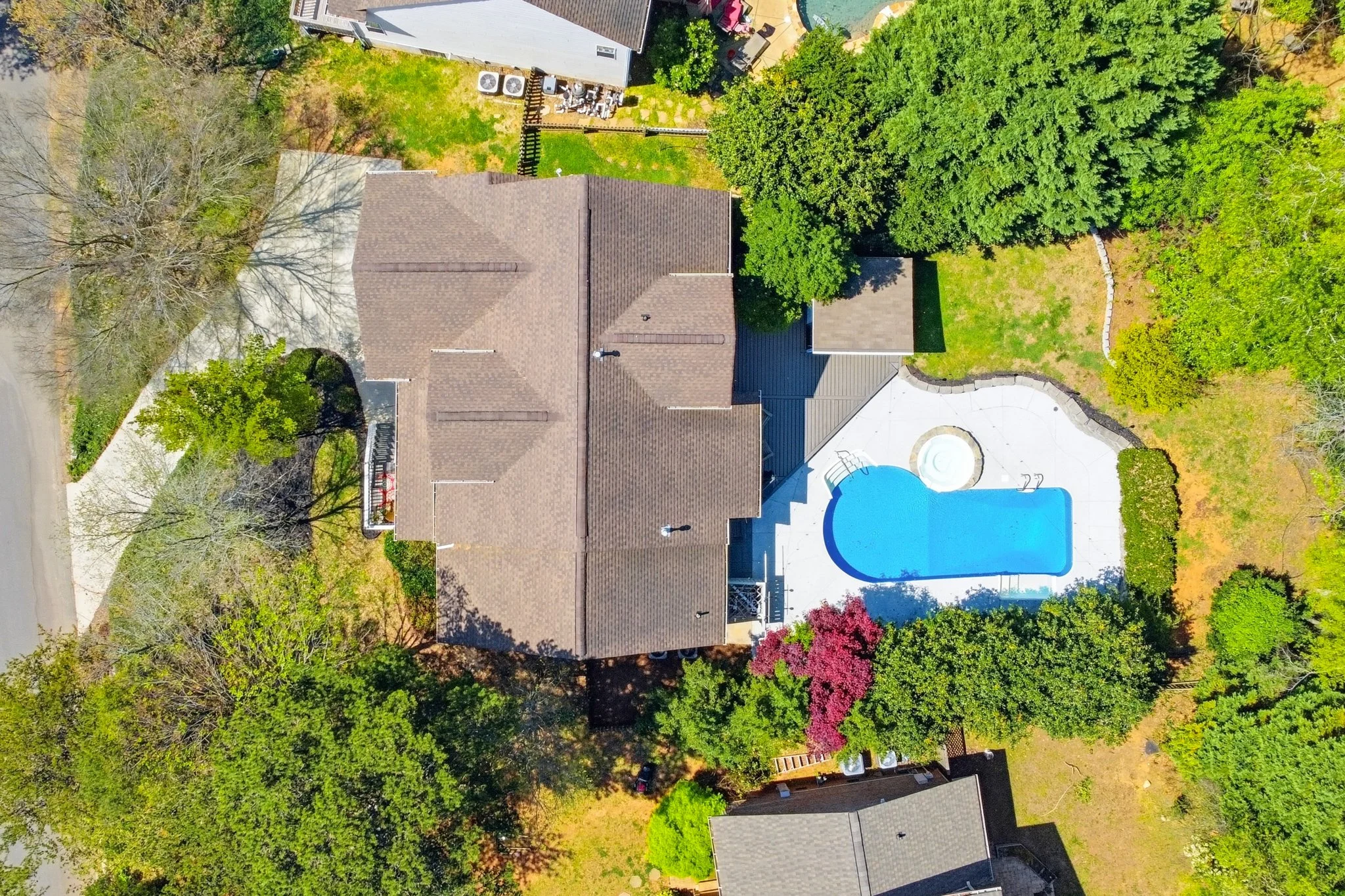 Aerial view of a house with a brown roof, surrounded by trees and green lawn. A backyard features a swimming pool with a curved shape and a white pool cover at one end.