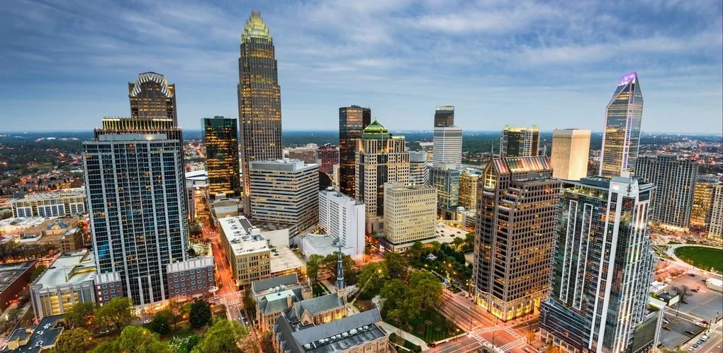 Aerial view of downtown Charlotte, North Carolina city skyline at dusk with illuminated skyscrapers and clouds overhead.