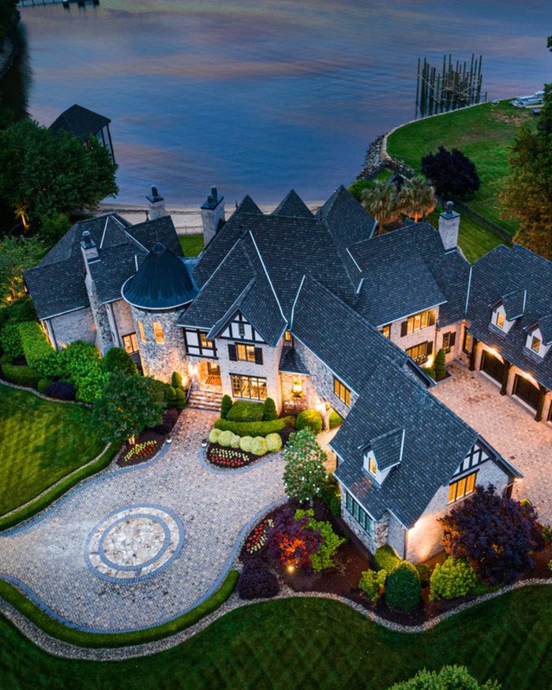 A large, elegant house by the water at dusk with illuminated windows, a landscaped garden, and a waterfront with docks.