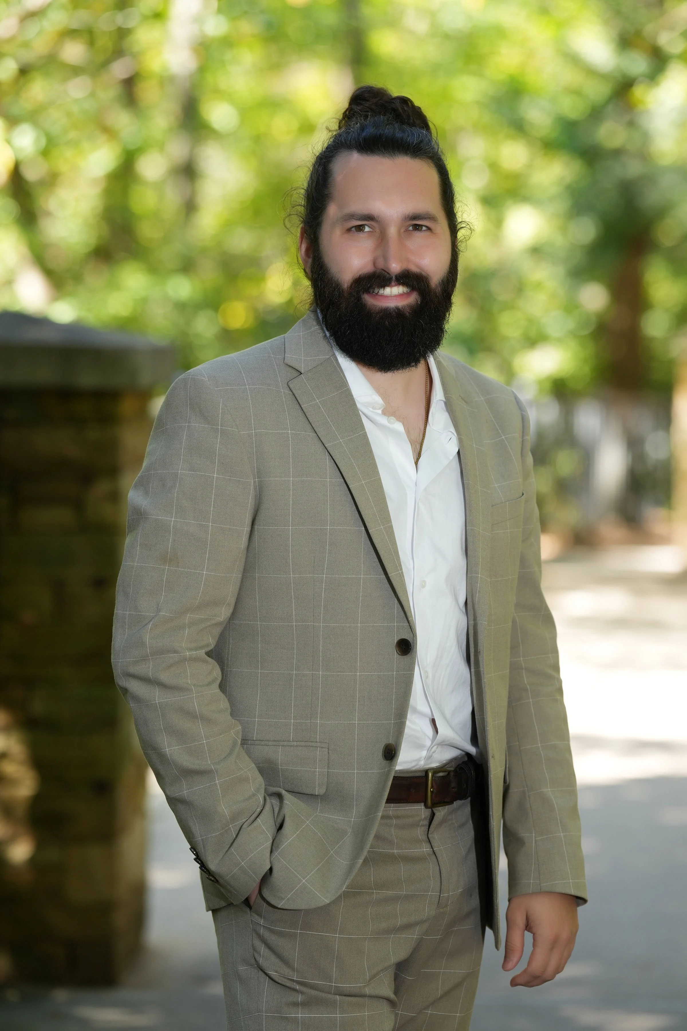 A man with a beard in a beige checkered suit, white shirt, and dark belt, standing outdoors with a blurred green forest background.
