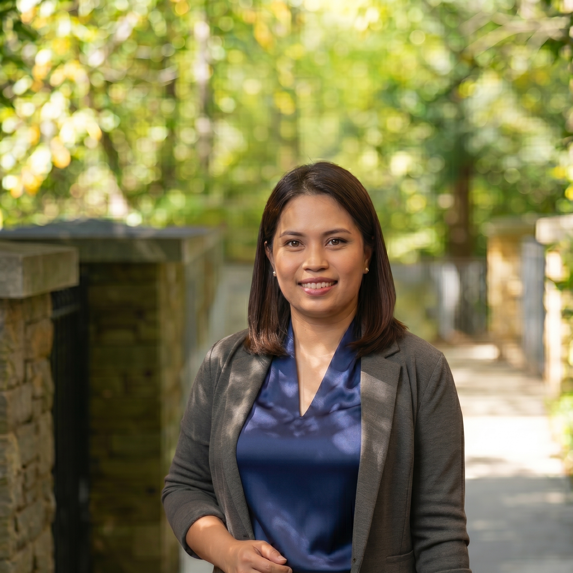 A woman with dark brown hair and a gray blazer wearing a blue blouse standing outside in front of a blurred green and yellow foliage background.