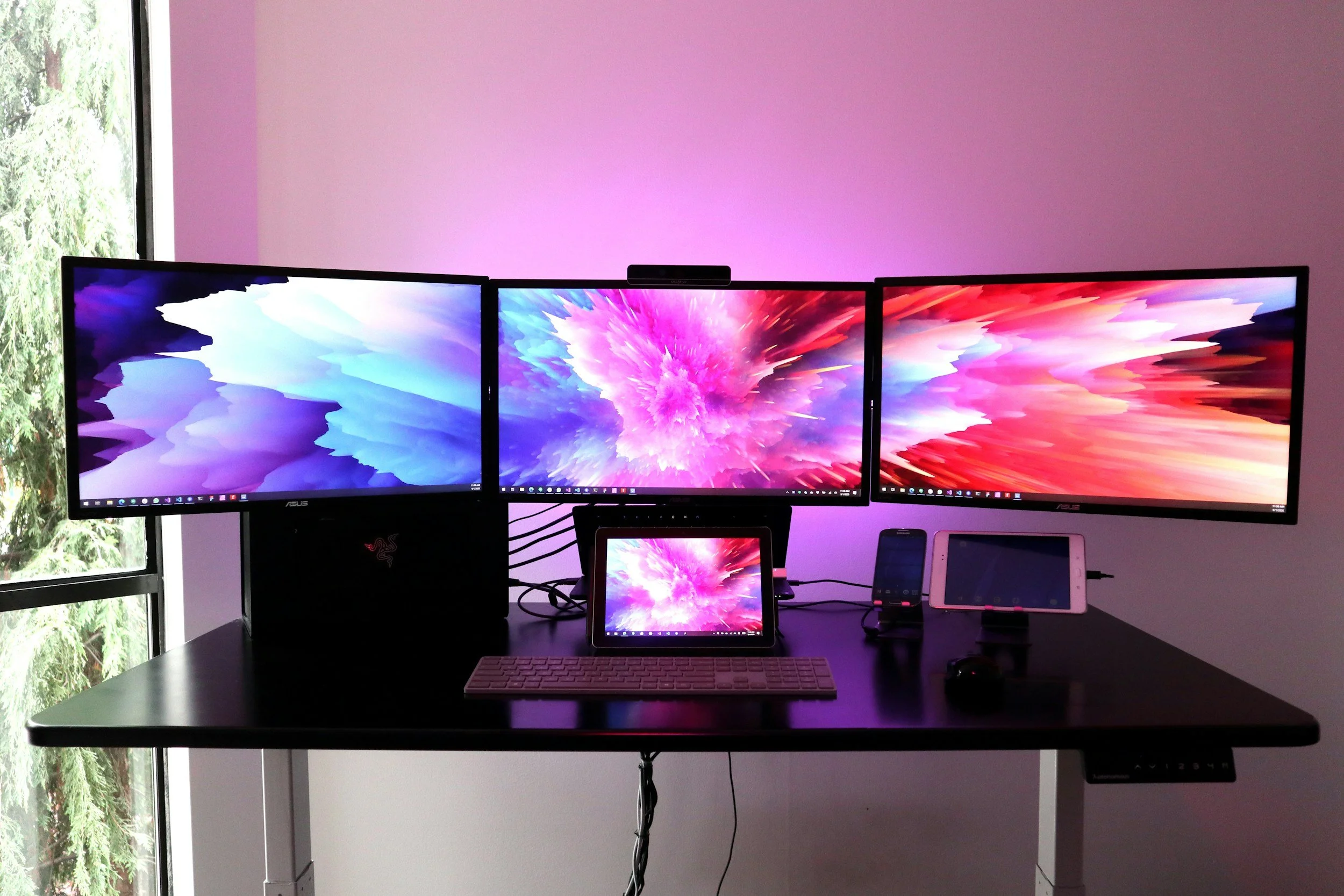 A computer workstation with three monitors displaying colorful abstract backgrounds, a tablet, a keyboard, and various cables on a black desk in a room with pink walls and a window showing green trees outside.