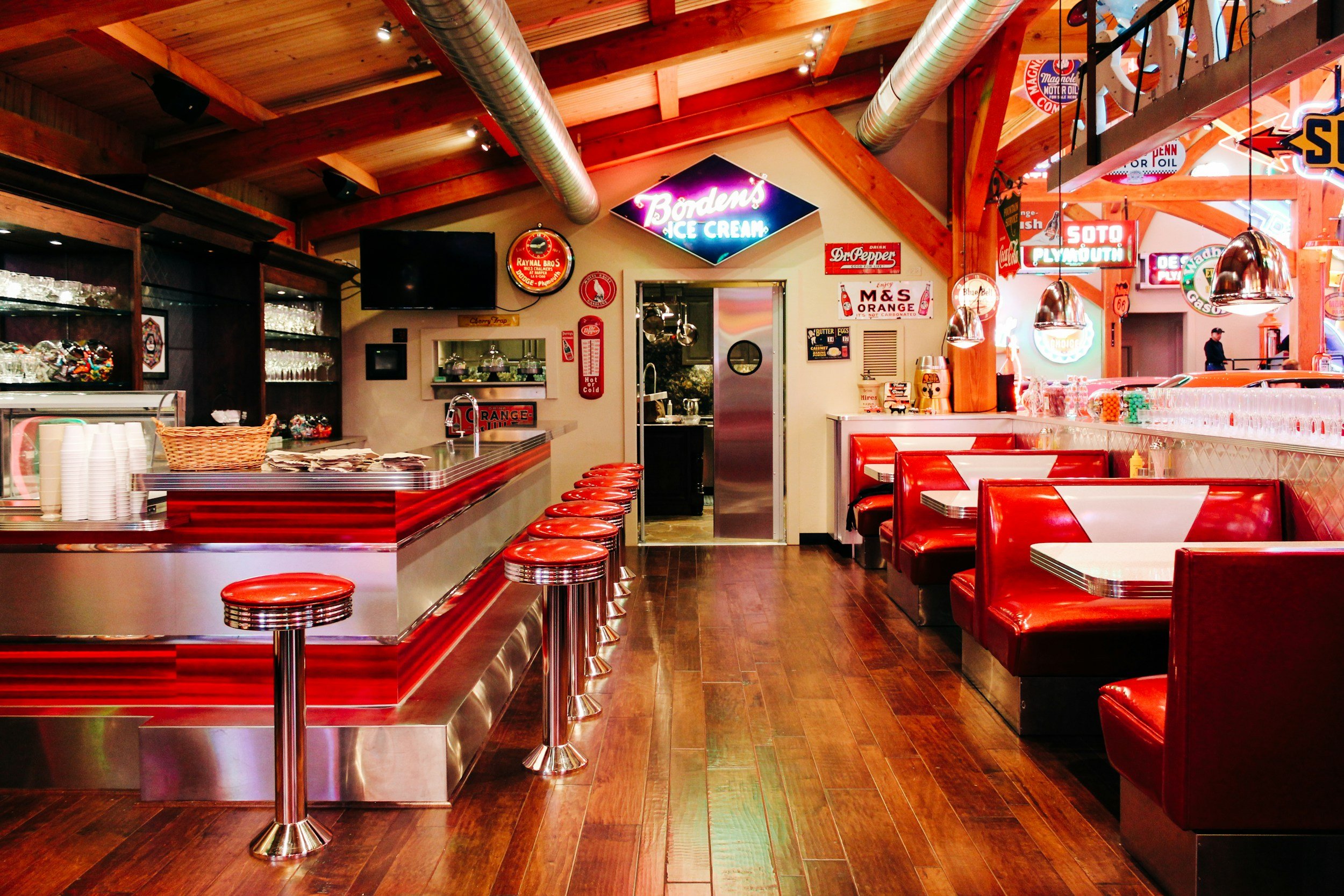 Interior of a vintage-style diner with red leather booths, chrome bar stools, wood ceiling, and neon signs on the walls.
