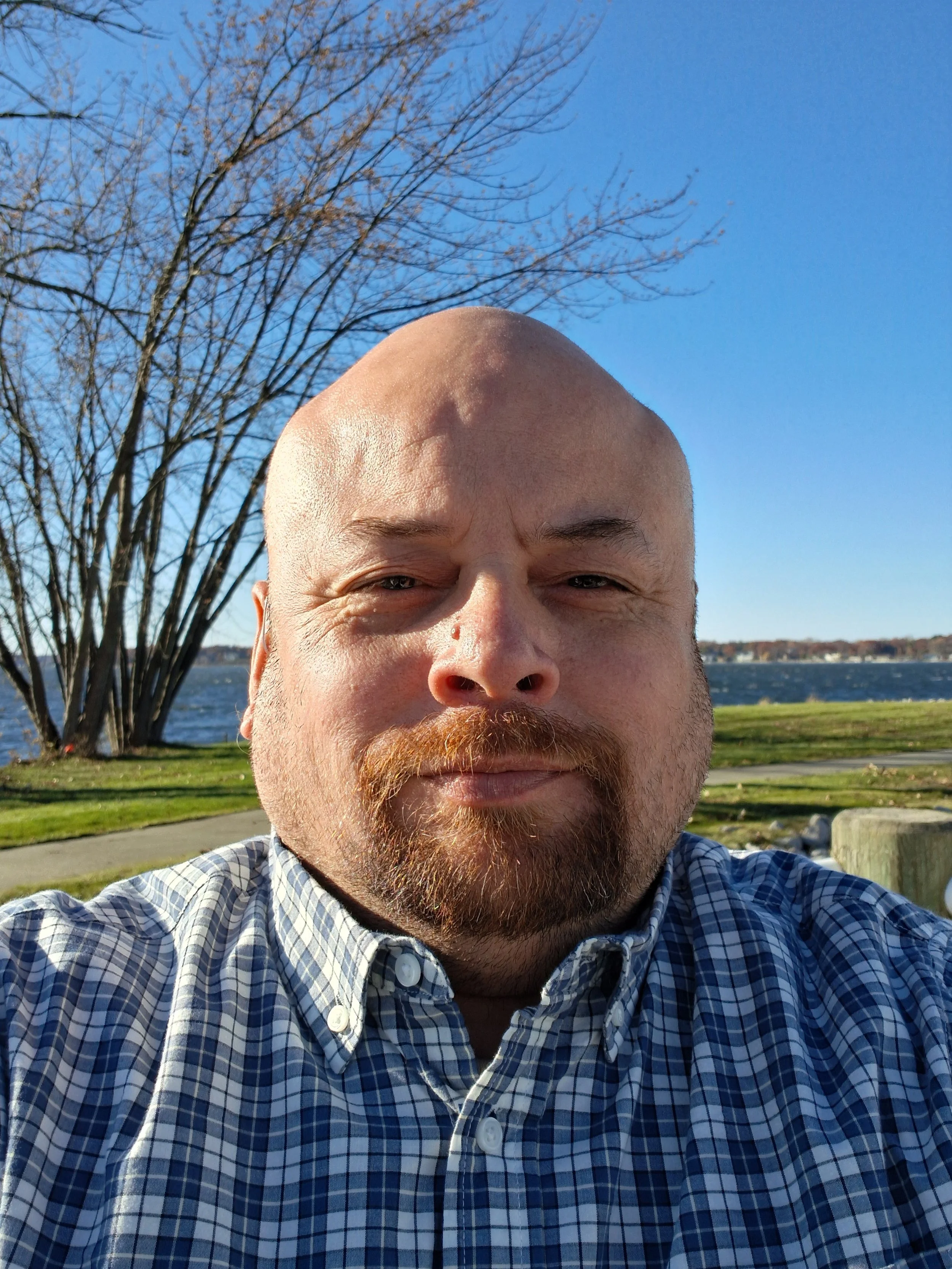 A man with a beard and mustache smiling outdoors in front of a tree with bare branches, near a body of water, on a sunny day.