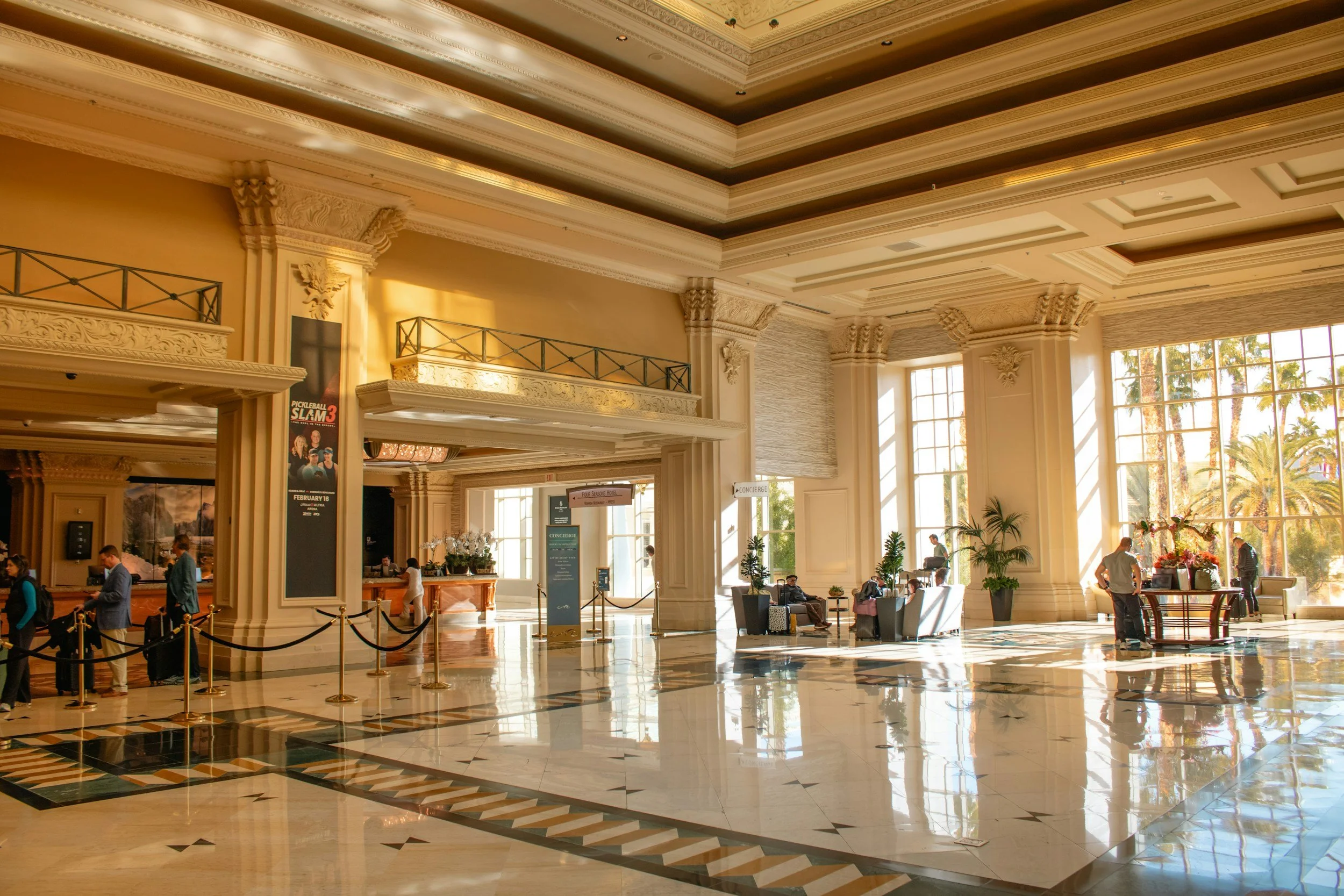 Interior of a grand hotel lobby with high ornate ceilings, large windows, and polished marble floors. There are people seated and standing, with some checking in at a reception desk, and decorative plants and furniture throughout.