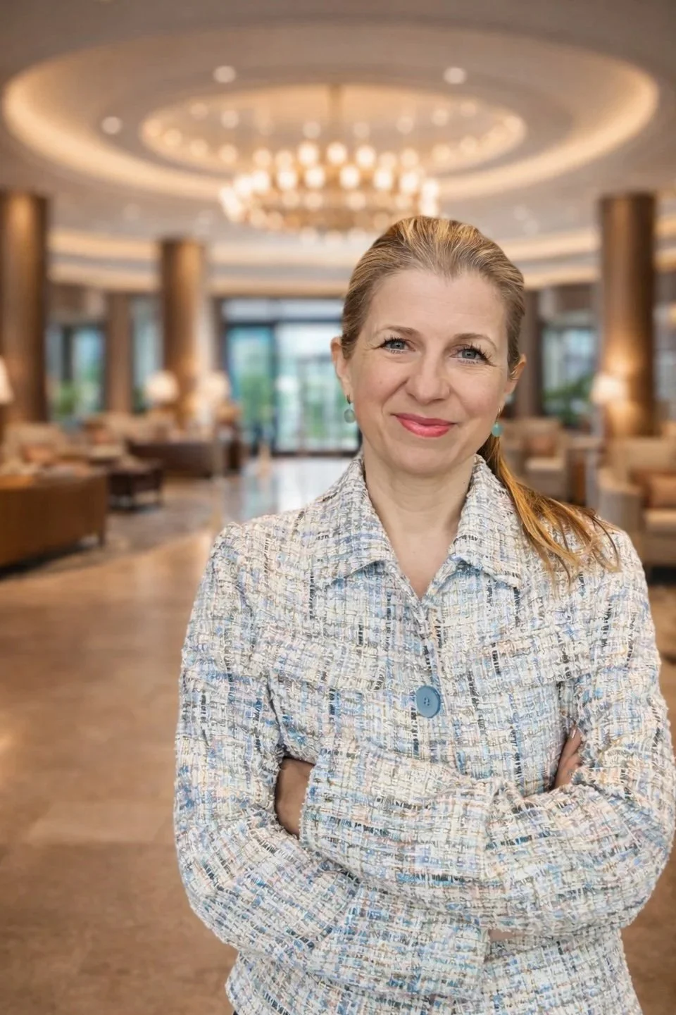 A woman with light brown hair tied back, smiling, standing with arms crossed in a hotel lobby with a chandelier and large windows in the background.