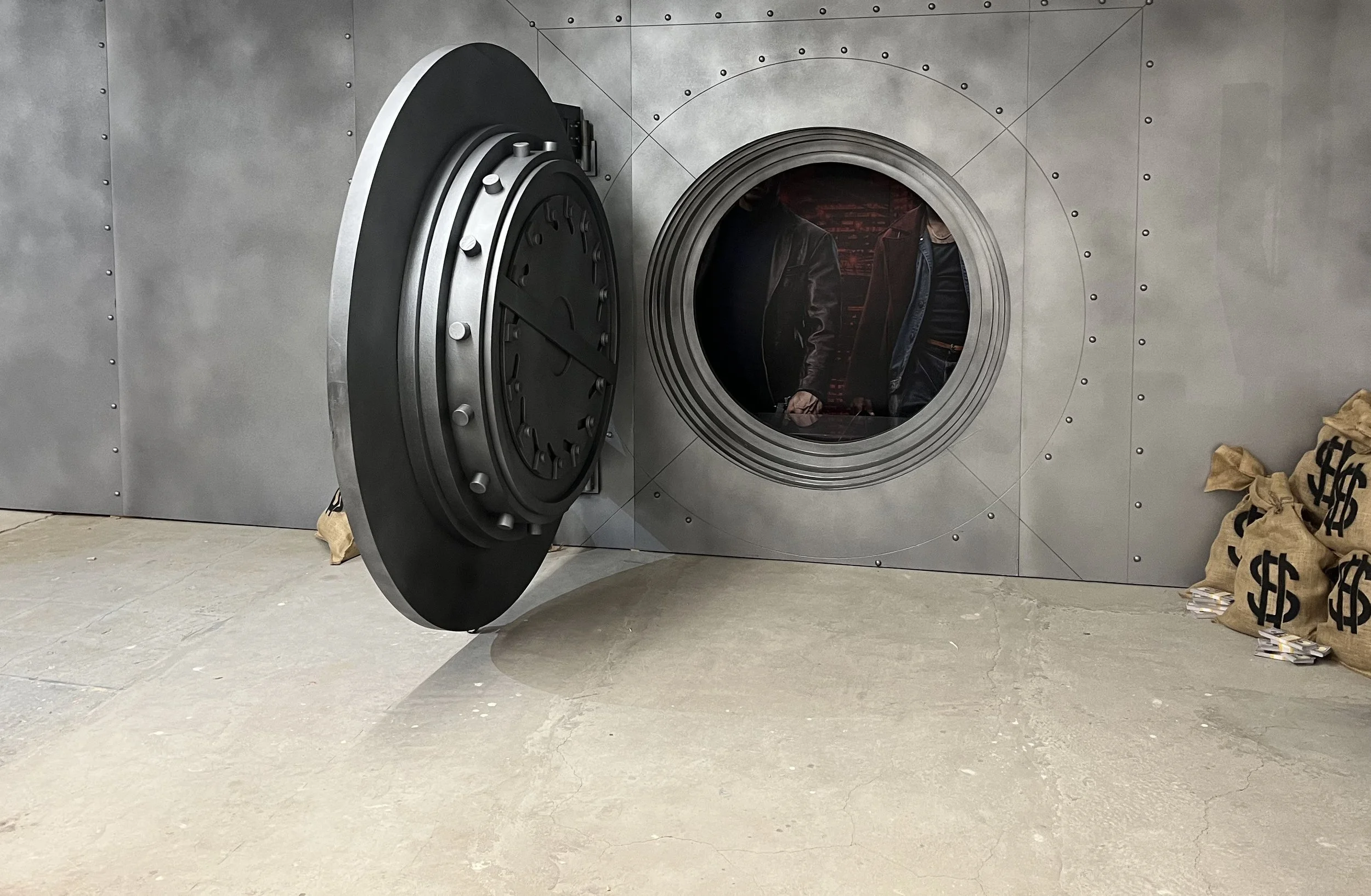 A gray metallic bank vault door is open, revealing a dark interior with two people inspecting the vault. There are sandbags with dollar signs on the ground to the right.
