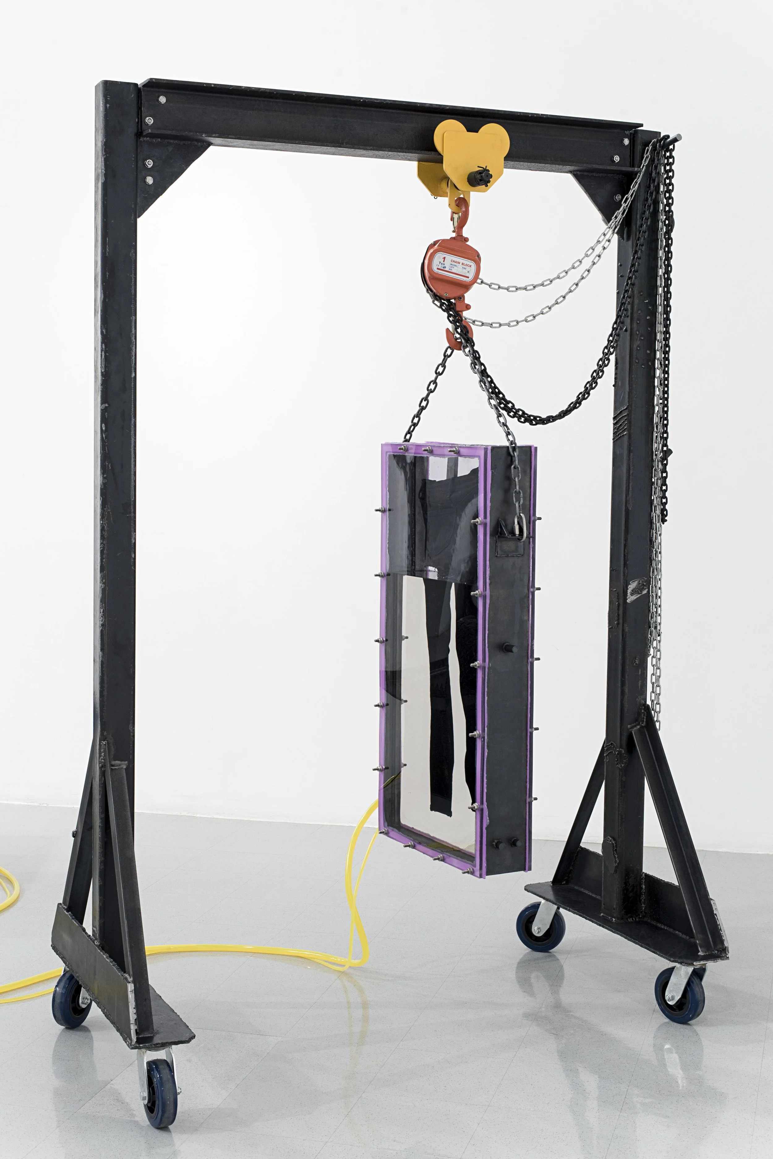 An industrial robot with a black metal frame on wheels, featuring chains and a yellow bear-shaped hook, holding a reflective rectangular glass panel with a purple border, in an art gallery.