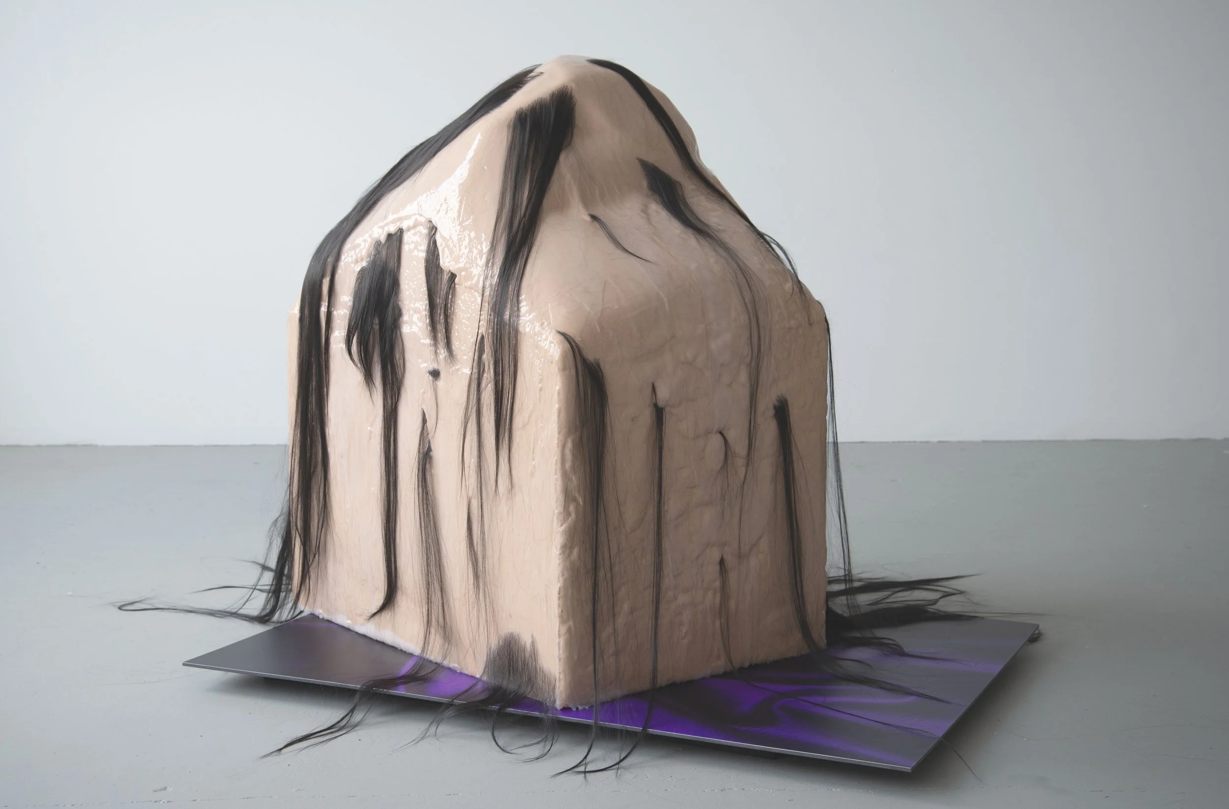 A sculptural art piece resembling a human head with long black hair, made from a light-colored material, possibly resin or plaster, with shiny and textured surfaces, placed on a gray platform in a minimalistic gallery space.