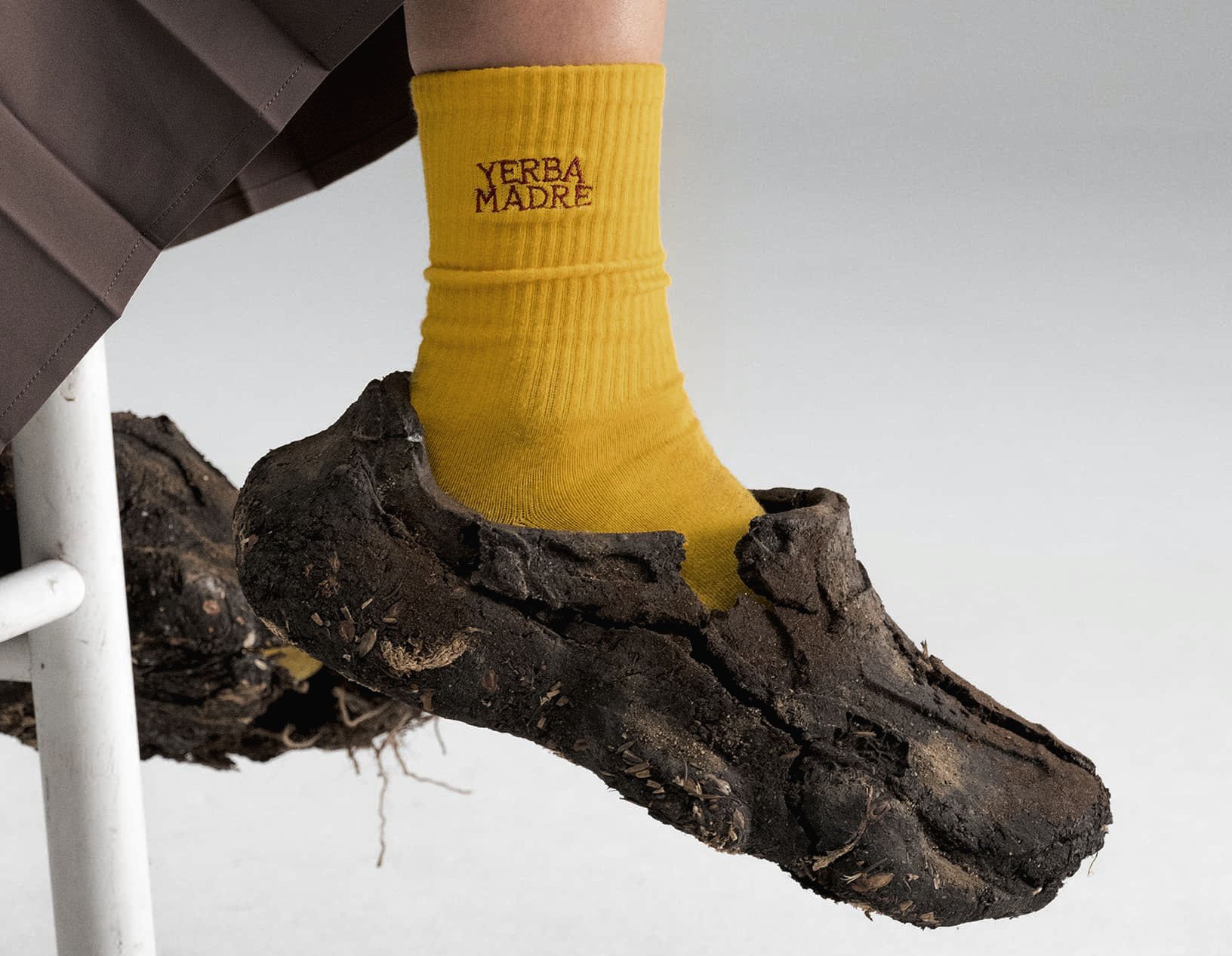 A yellow sock with red text that reads 'YERBA MADRE' is worn on a foot, which is stepping on a piece of rough, dark wood or bark, against a plain light background.