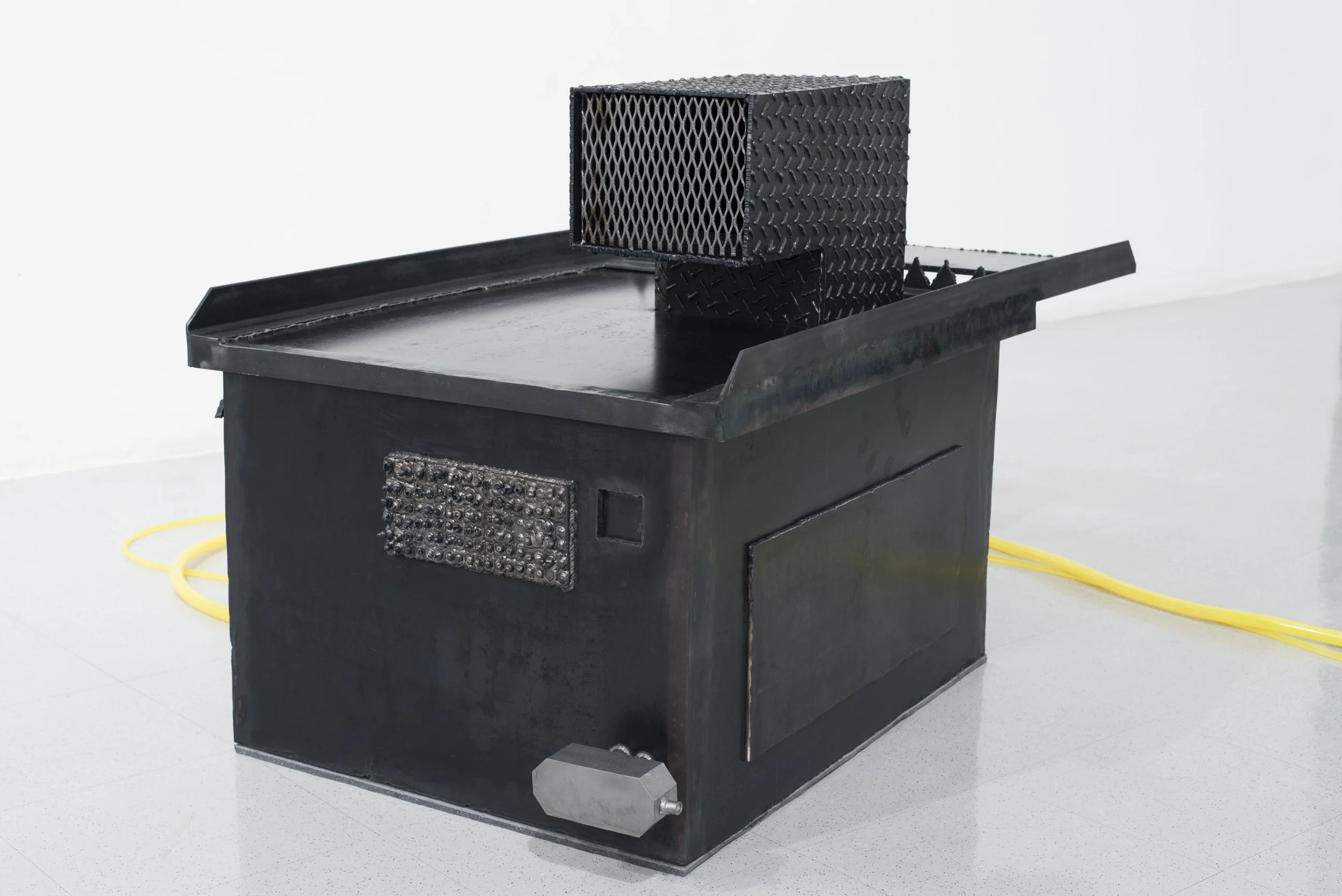 Black industrial oven or furnace on a platform with a grated metal box on top, connected to yellow electrical cords in a white-walled room.