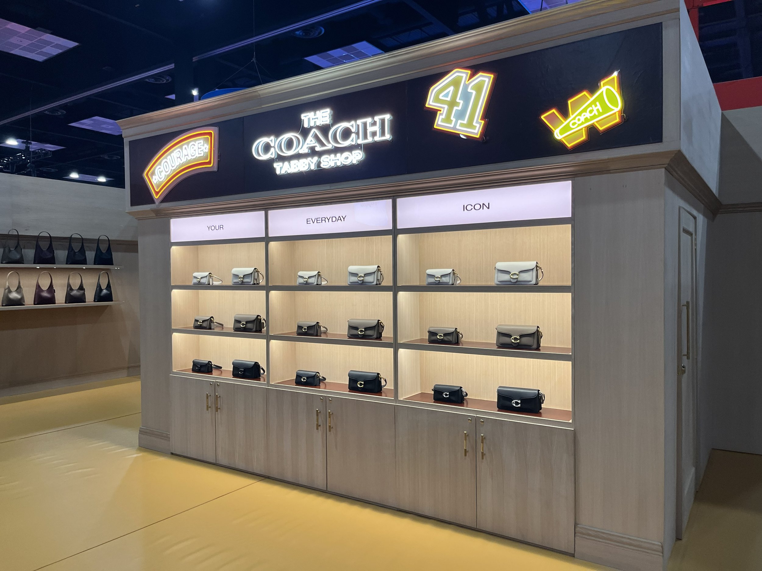 Store display with handbags arranged on illuminated shelves, sign shows 'The Coach Tabby Shop' with neon signs reading 'Courage,' '$1,' and a yellow neon 'Coach' logo.