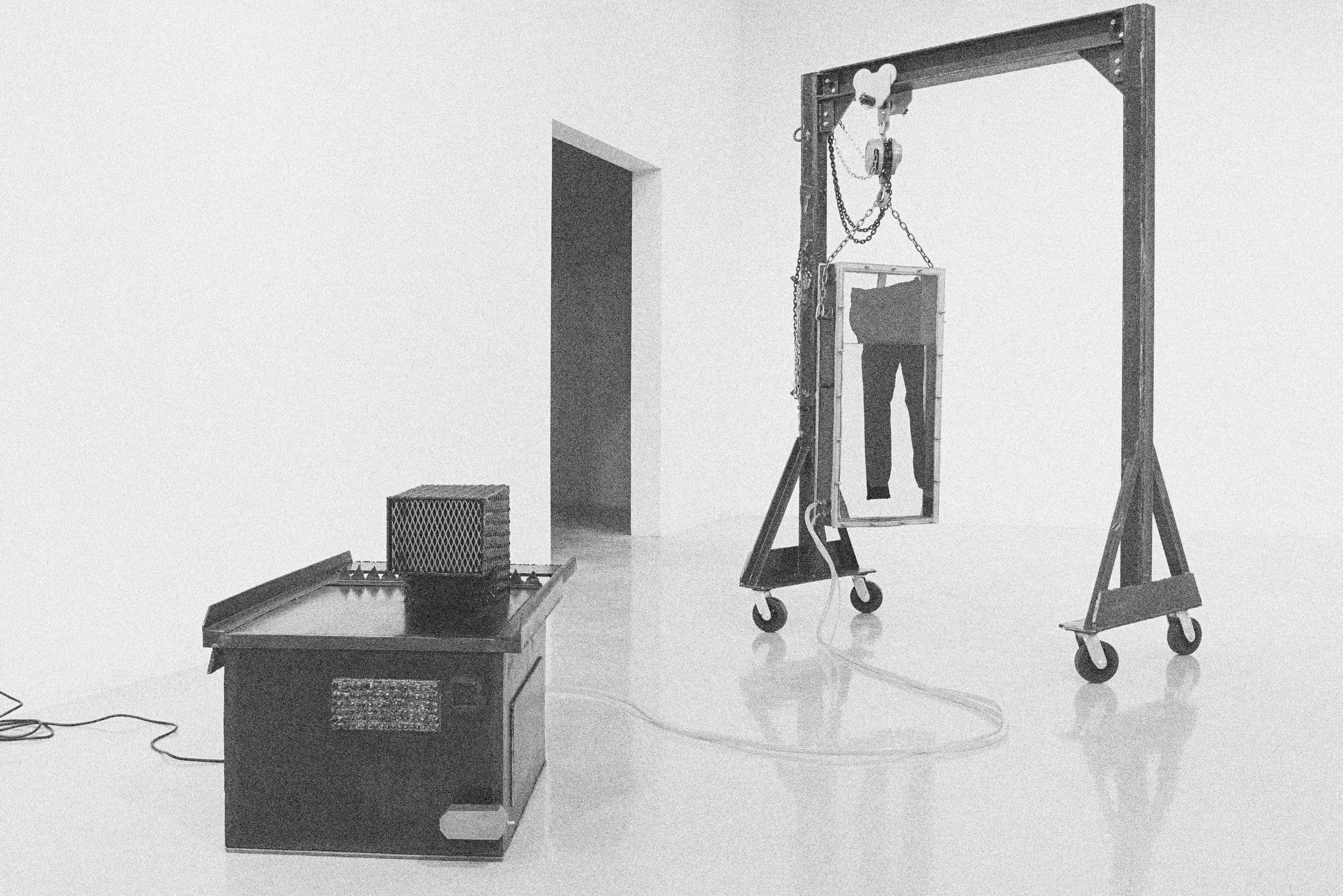 A series of metal chambers in a white room with one chamber hanging and filled with a pair of black jeans.