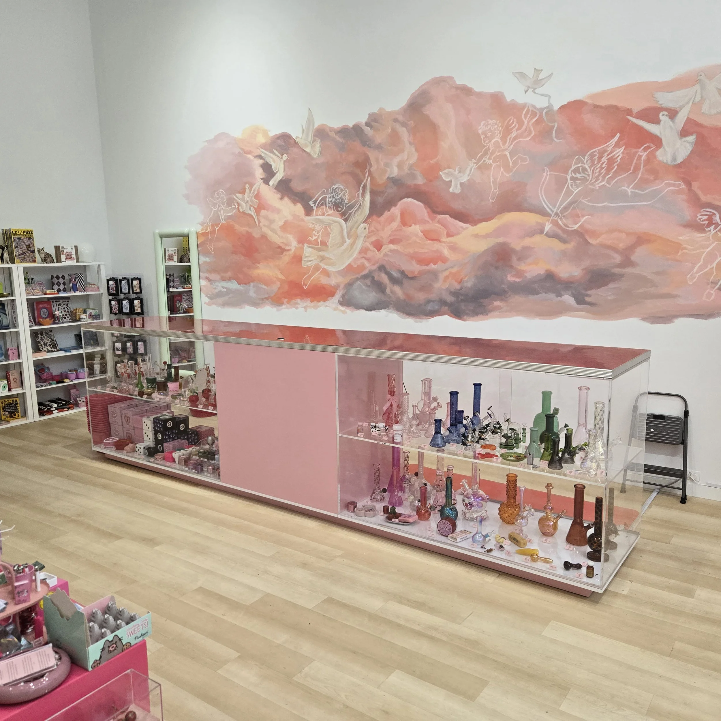 Display of various pink-themed glassware and accessories in a store with a pink partition and a cloudy sky mural with cherubs on the wall.