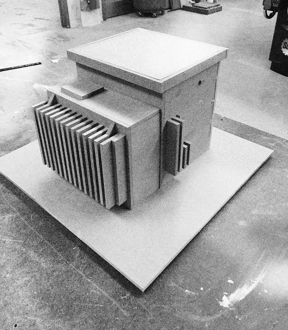 A model of a small building or structure with modern design, featuring vertical slats, on a white platform in a workshop or storage area.