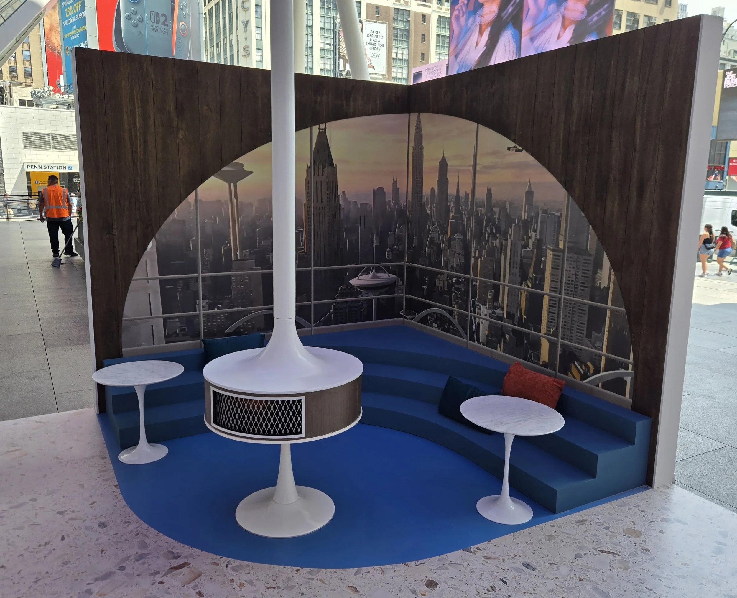 Modern indoor seating area with blue carpet, built-in bench with red and blue cushions, white side tables, and a backdrop featuring a cityscape of New York City skyline with Empire State Building and Chrysler Building at sunset.