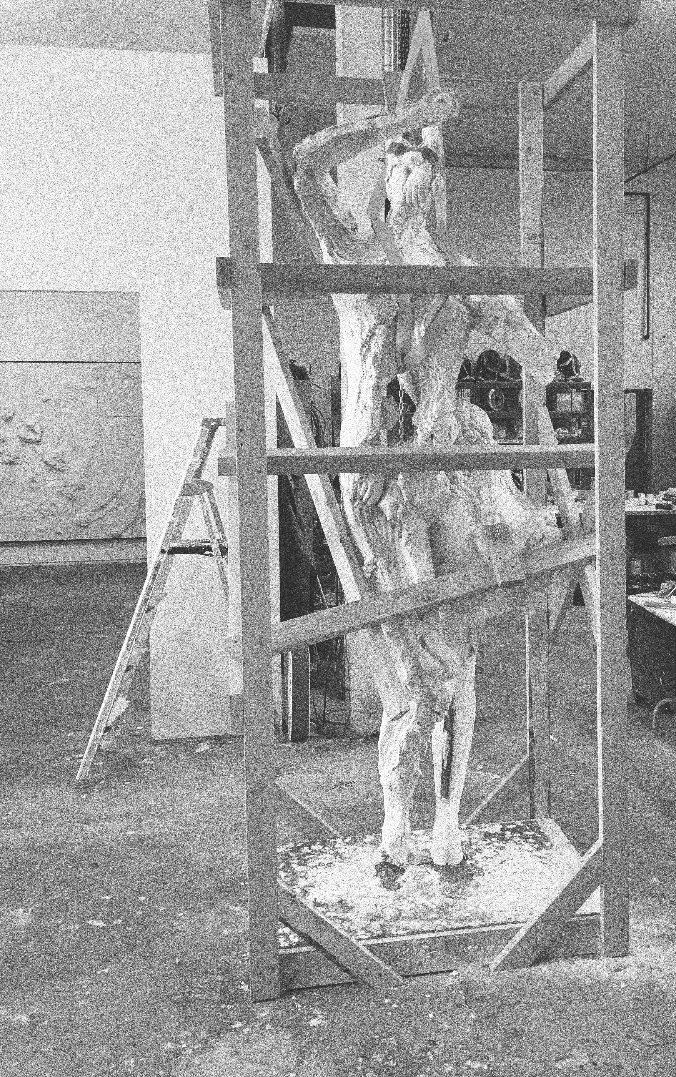 A sculpture of a human figure, possibly an angel or mythological person, within a wooden frame and metal scaffolding. The sculpture appears to be in progress, with an abstract or unfinished form, in an art studio or workshop setting.