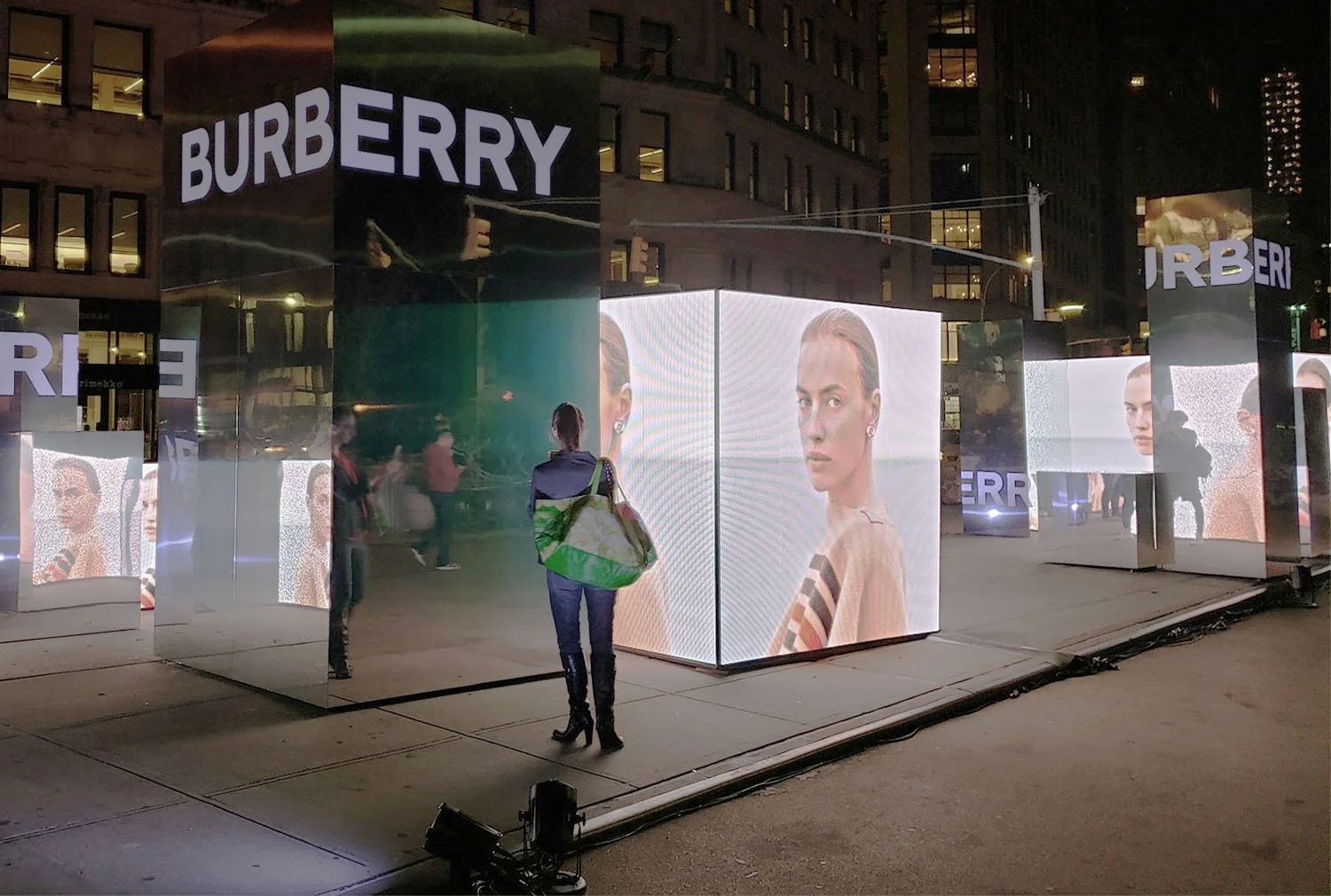 Nighttime outdoor installation with illuminated digital screens displaying fashion model images and the word "BURBERRY" in large letters at the top of reflective display panels, with a person standing and looking at the screens.